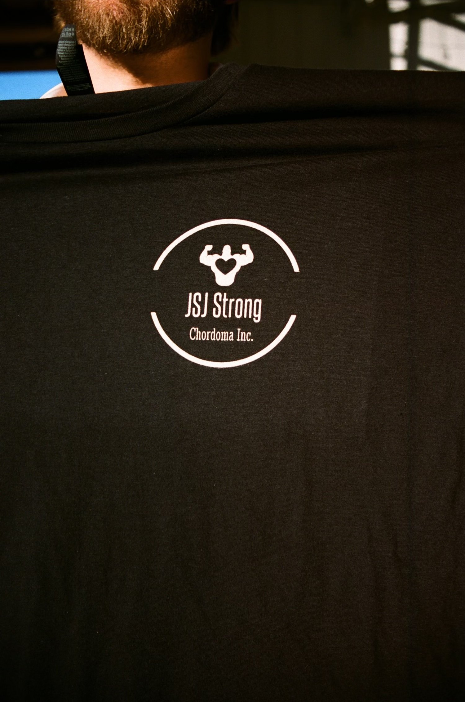 Black t-shirt with "JSJ Strong Chordoma Inc." white logo featuring a flexed arm with a heart symbol.
