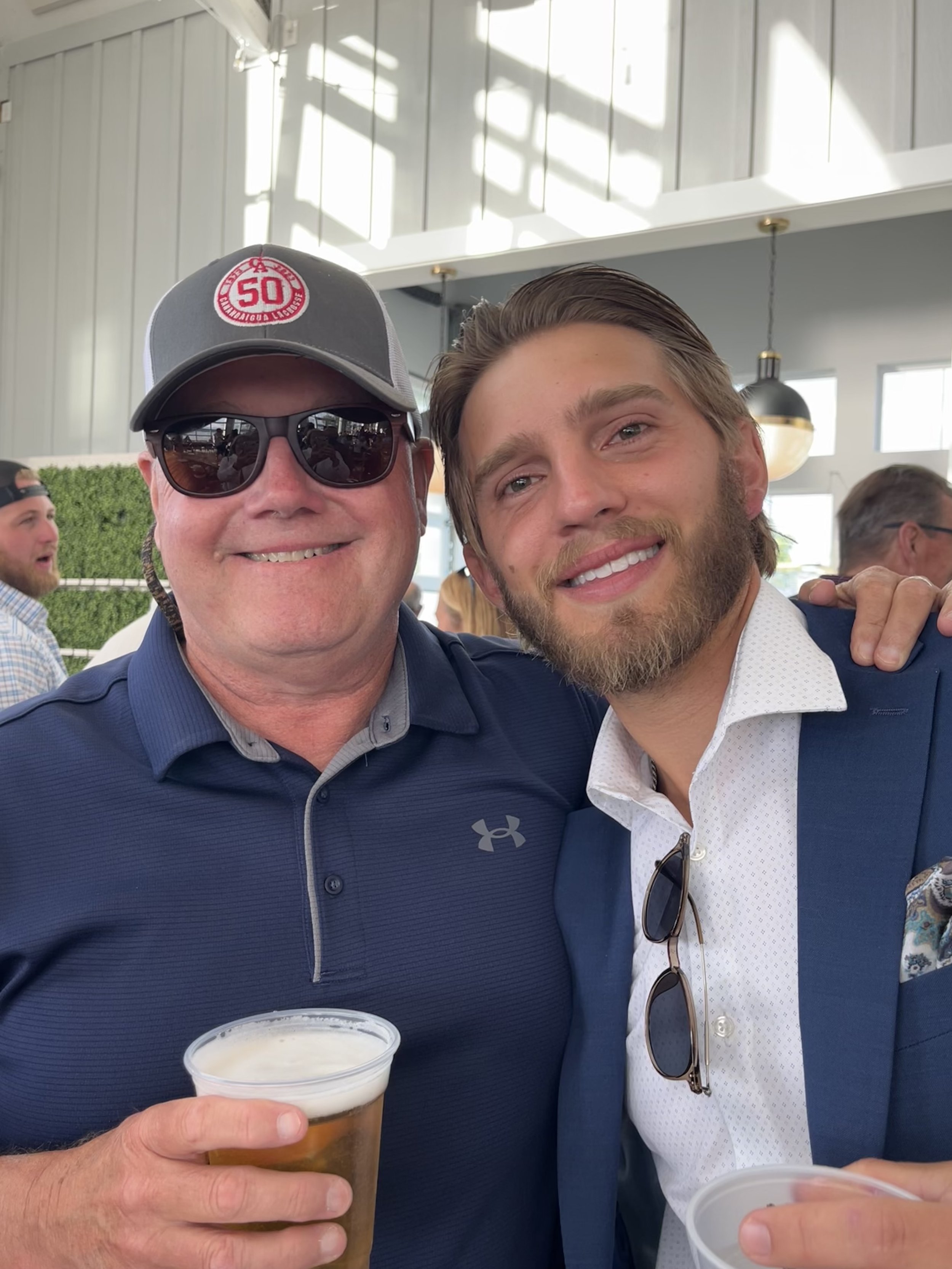 Two men smiling, one wearing a cap and sunglasses, holding a beer, the other in a suit with sunglasses and beard, indoors at an event.