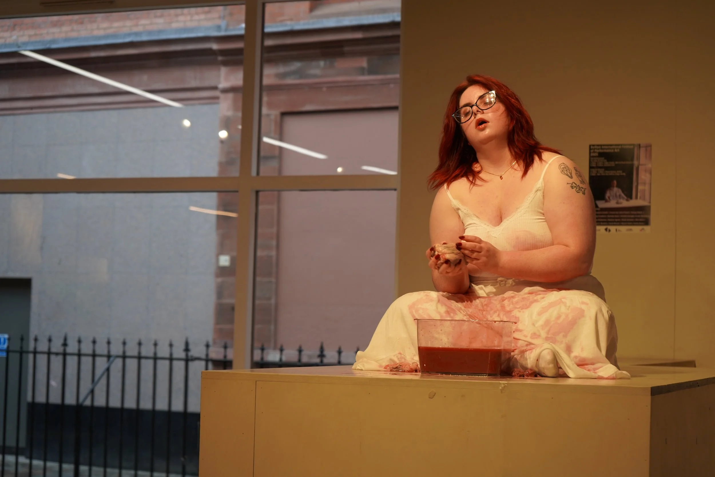 female in white dress and long red hair sitting cross legger on a high platform with a glass bowl half full of liquid in front of her. the front of the white dress is covered in blood