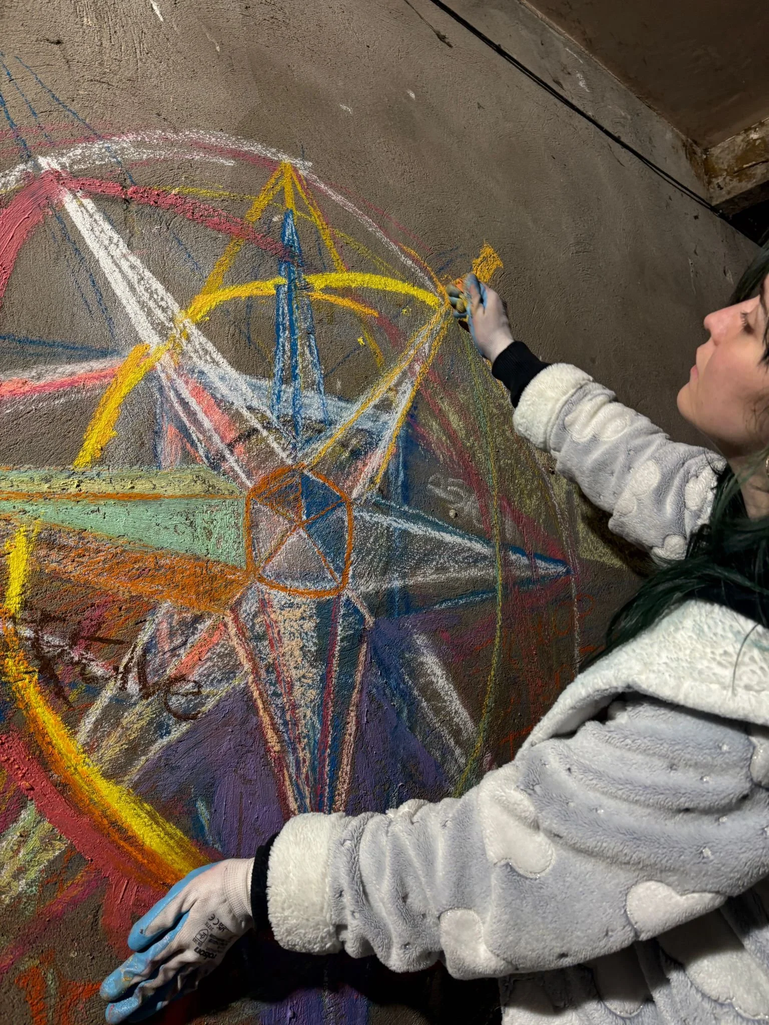 person leaning on the floor wearing a grey top, wearing gloves and drawing a multi-coloured eight pointed star surrounded by circles.