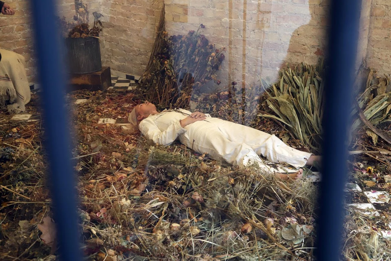 woman in white lying in a whited bricked space with the floor covered in dried grasses and planrs, Hnads clasped acros her belly as she lies flat on her back.