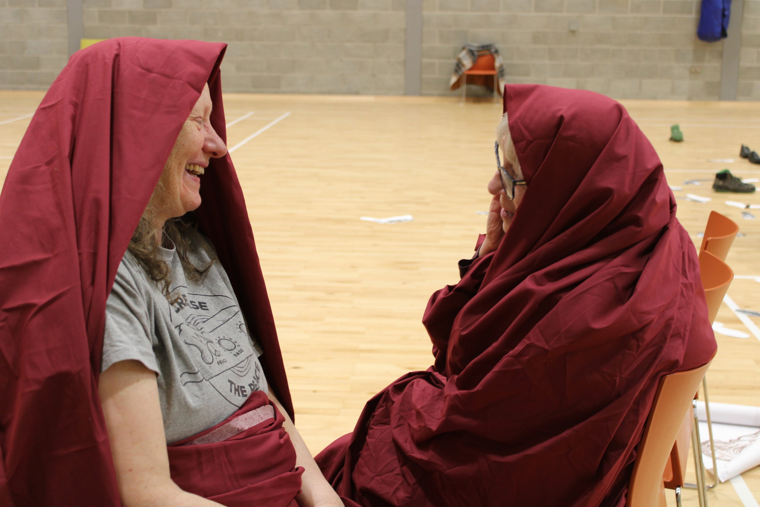 close up of two women swathed in red fabric  sitting on chairs facing each other, laughing