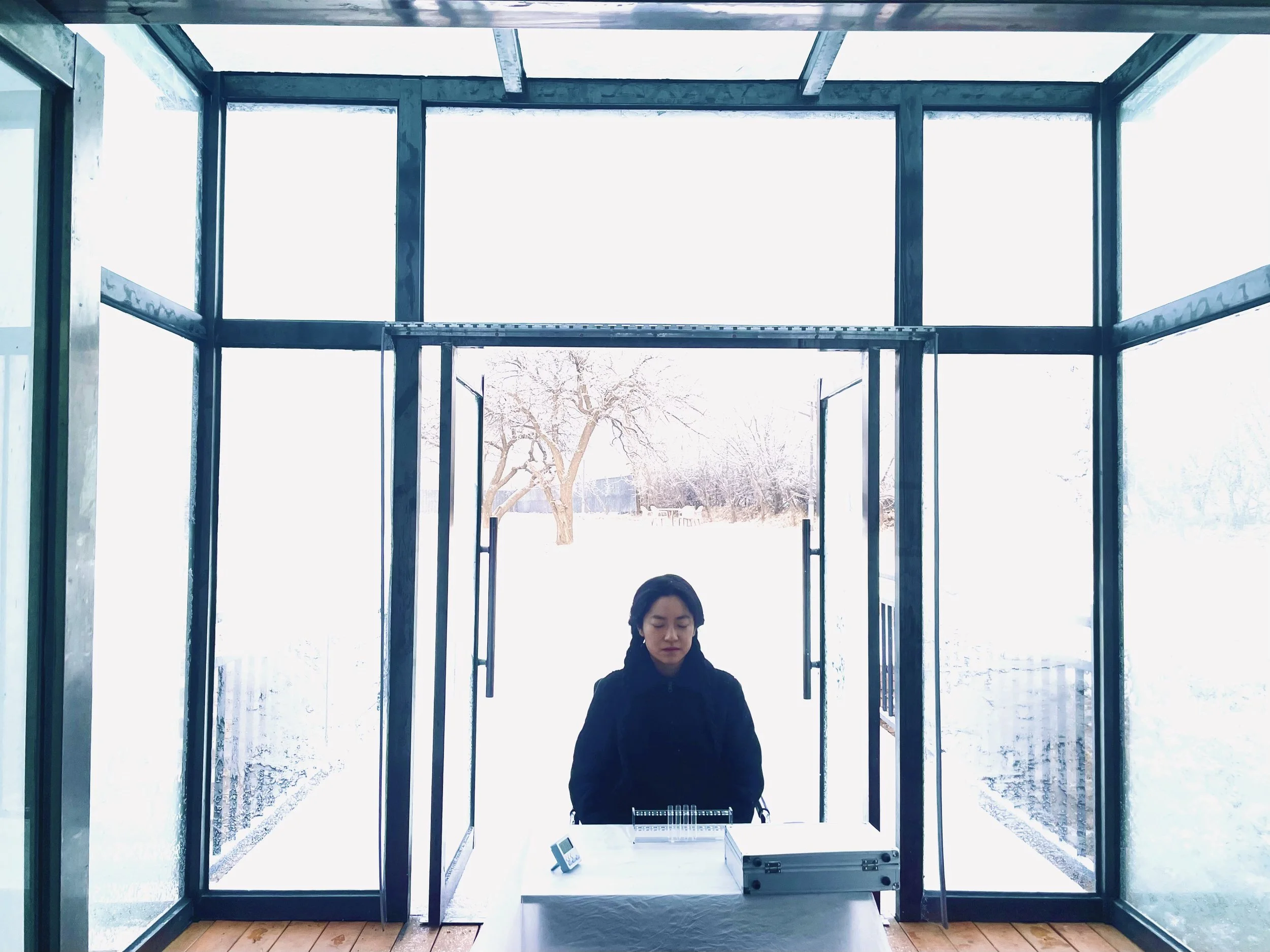 a female kneeling at a low table with small objects on the table. She wears black and has black hair tied back. In the background are open glass doors and floor to ceiling windows with a snowy treelined lanscape