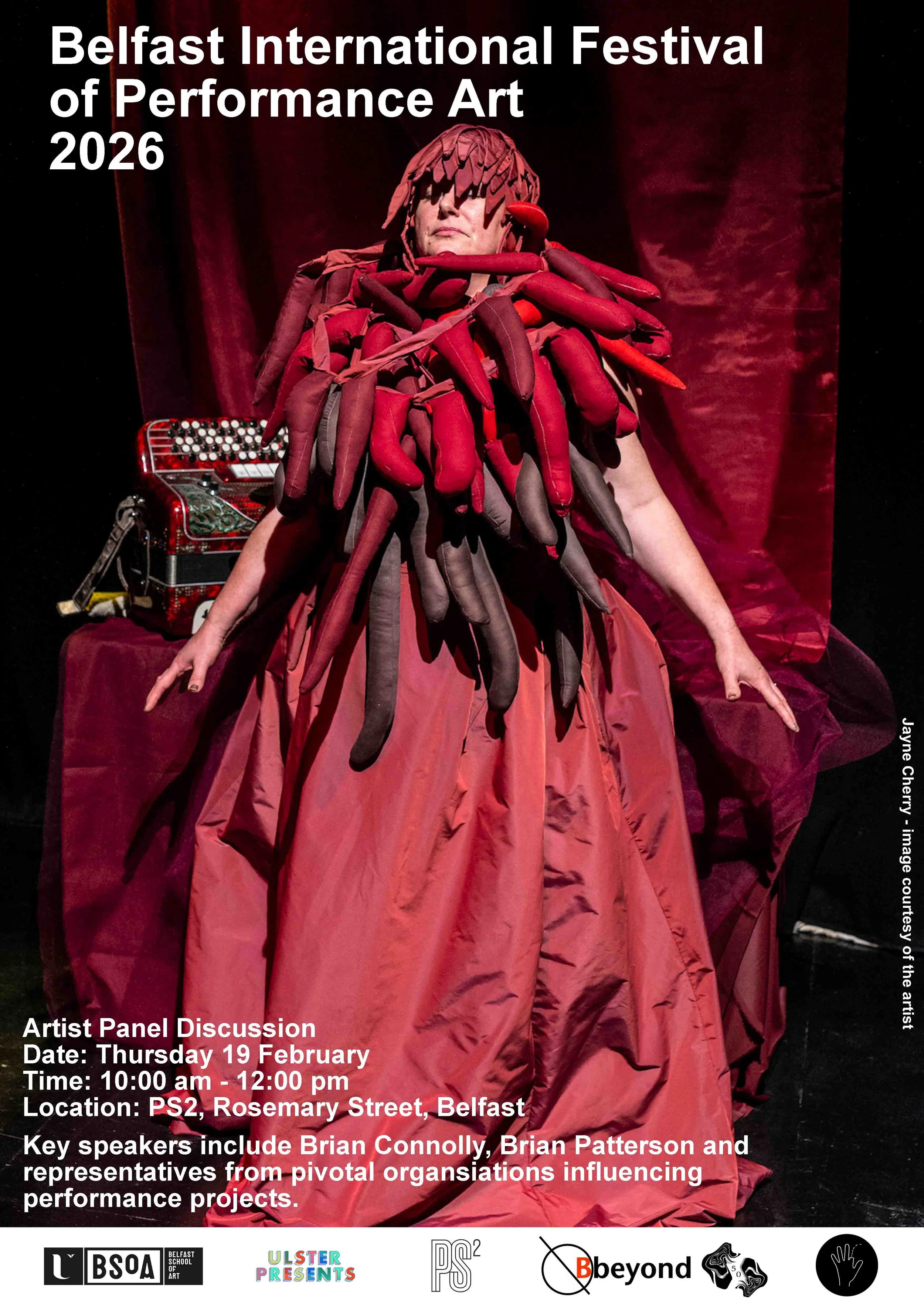 woman in a red dress, covered in red carrot sized and shaped fabric objects, arms outstretched waering a red texturedhat, red accordian on a table behind her and red curtain in background