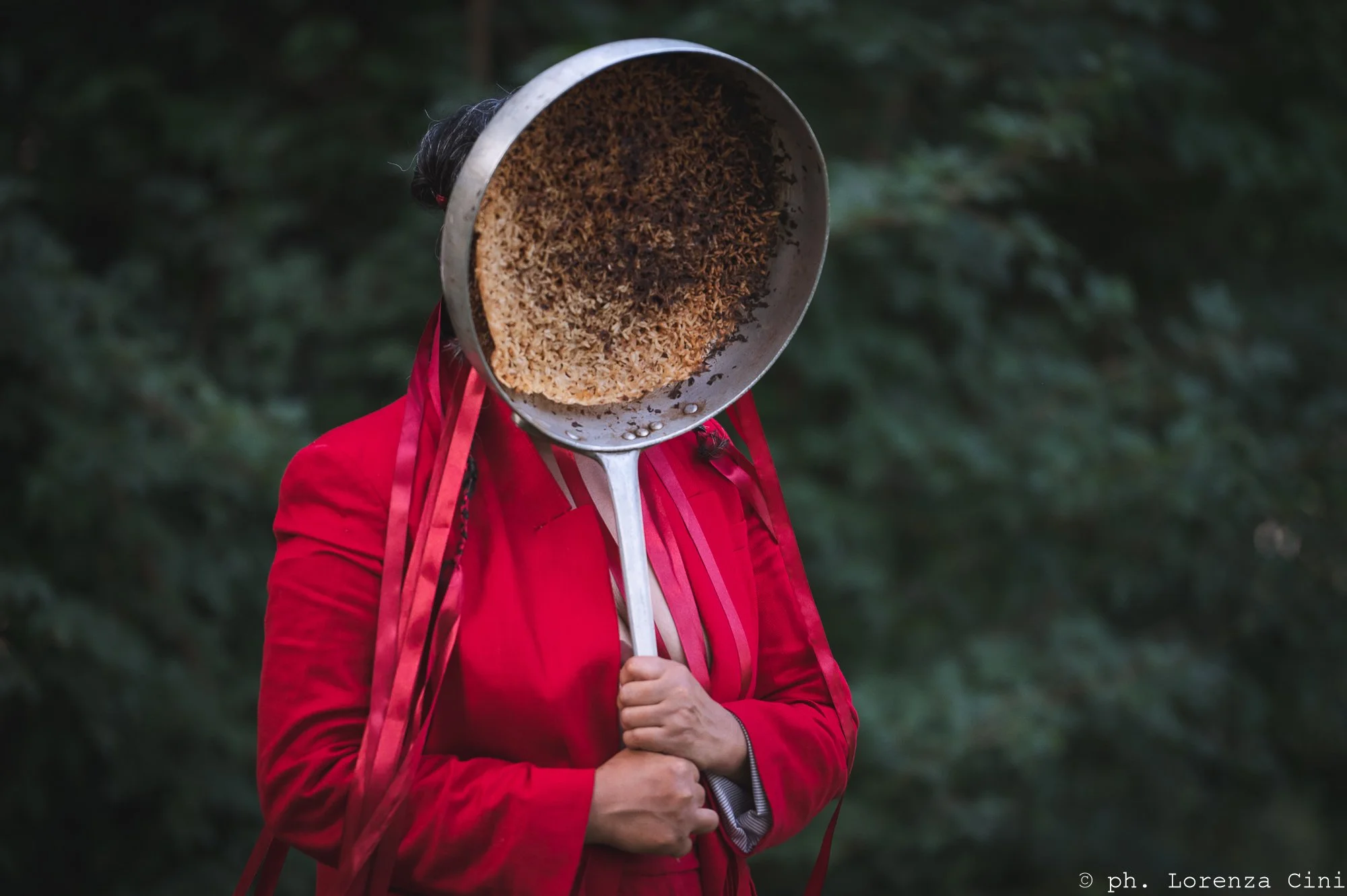 Female presenting person wearing bright red coat holding a burnt frying pan in front of their face with red ribbons flowing down their body