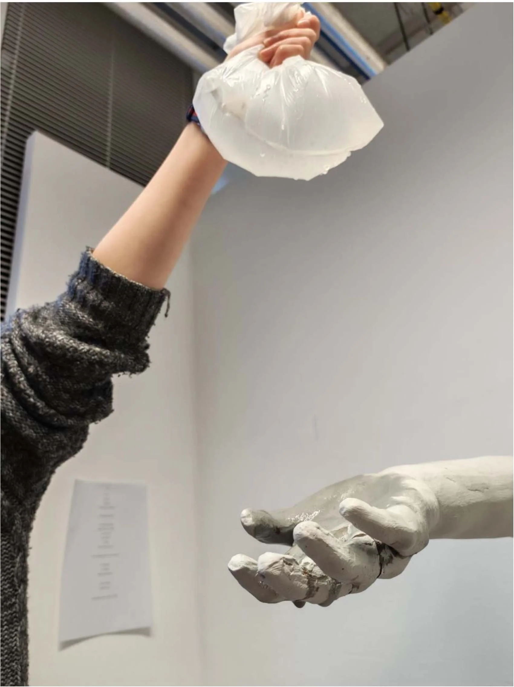 an arm stretches out holding a plastic bag full of water over a clay hand, reaching out as if to catch it.