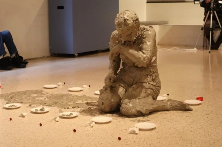 a figure kneeling on the floor completely covered in runny lay surrounded by small white plates and clay smeared on the floor.