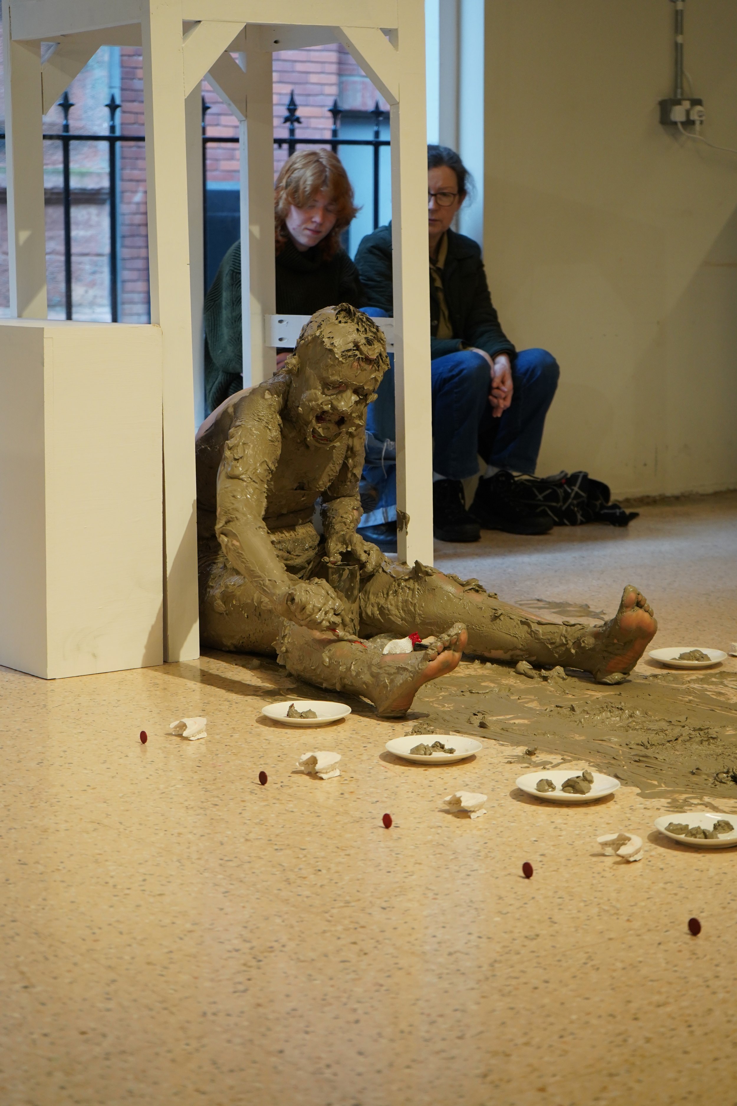 Person sitting on a floor with legs outstretched completely covered in muddy clay, legs outstretched. To their side is a row or white plates and small blobs of clay. Behind them is a white wooden arch.