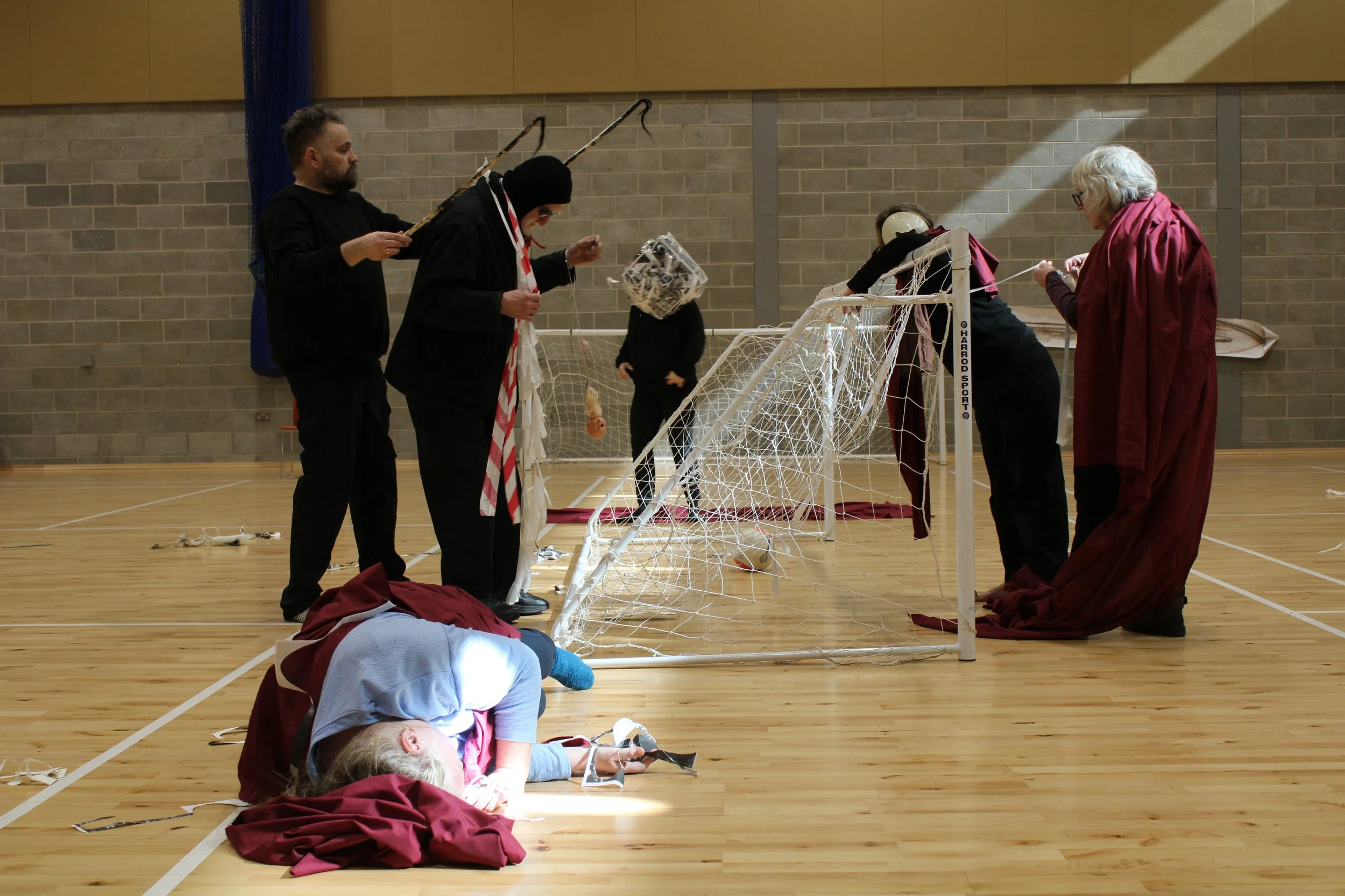 shaft of light on person swathed in red fabric lying on floor, people standing around  indoor football nets holding different objects, background people with paper covering their heads