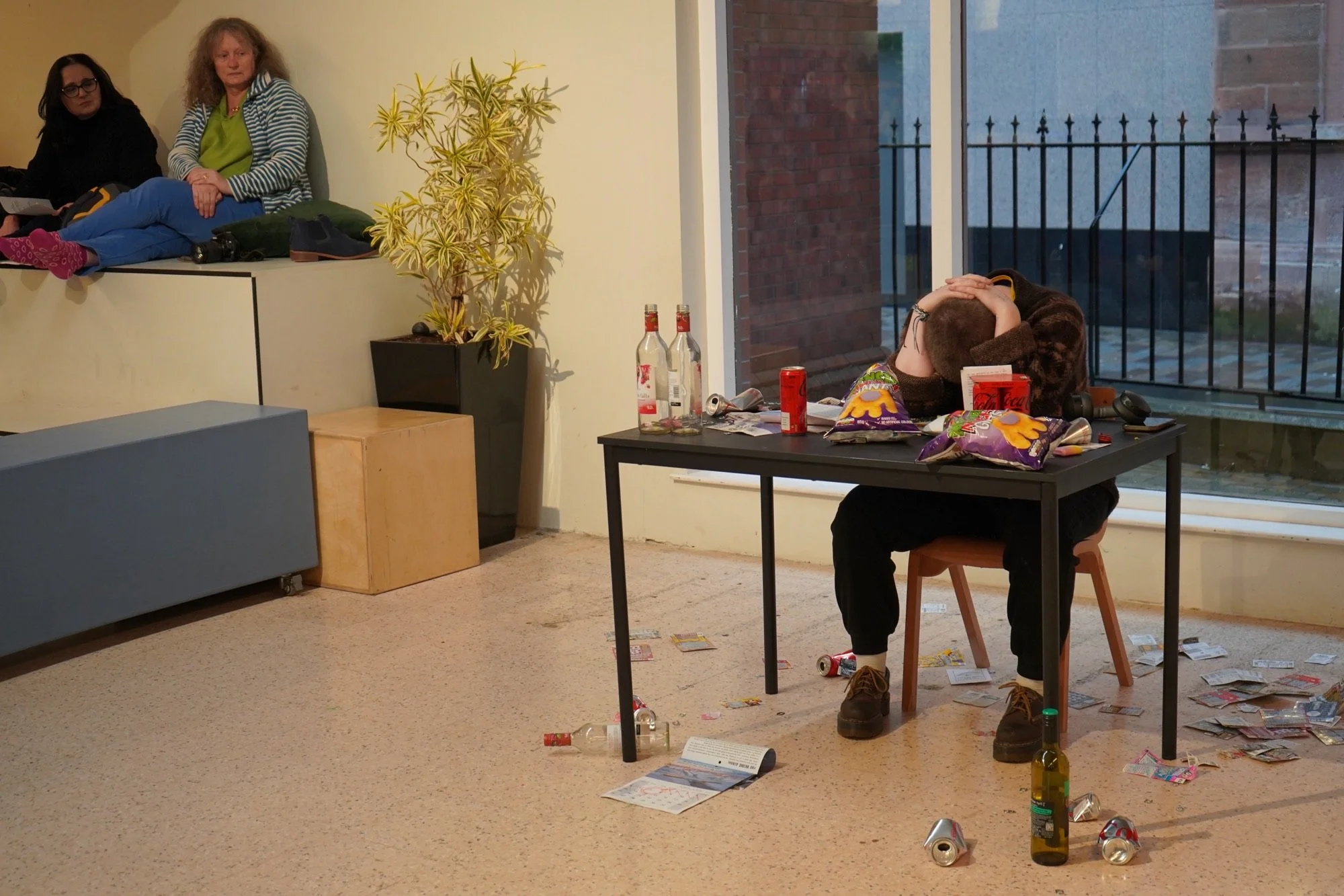 A person sitting at a desk covered in food wrappers, drink cans an bottles with their head on the table and hands clasped behind their head. They ware wearing dark clothes. There is a window behind them and two people on the left watching them