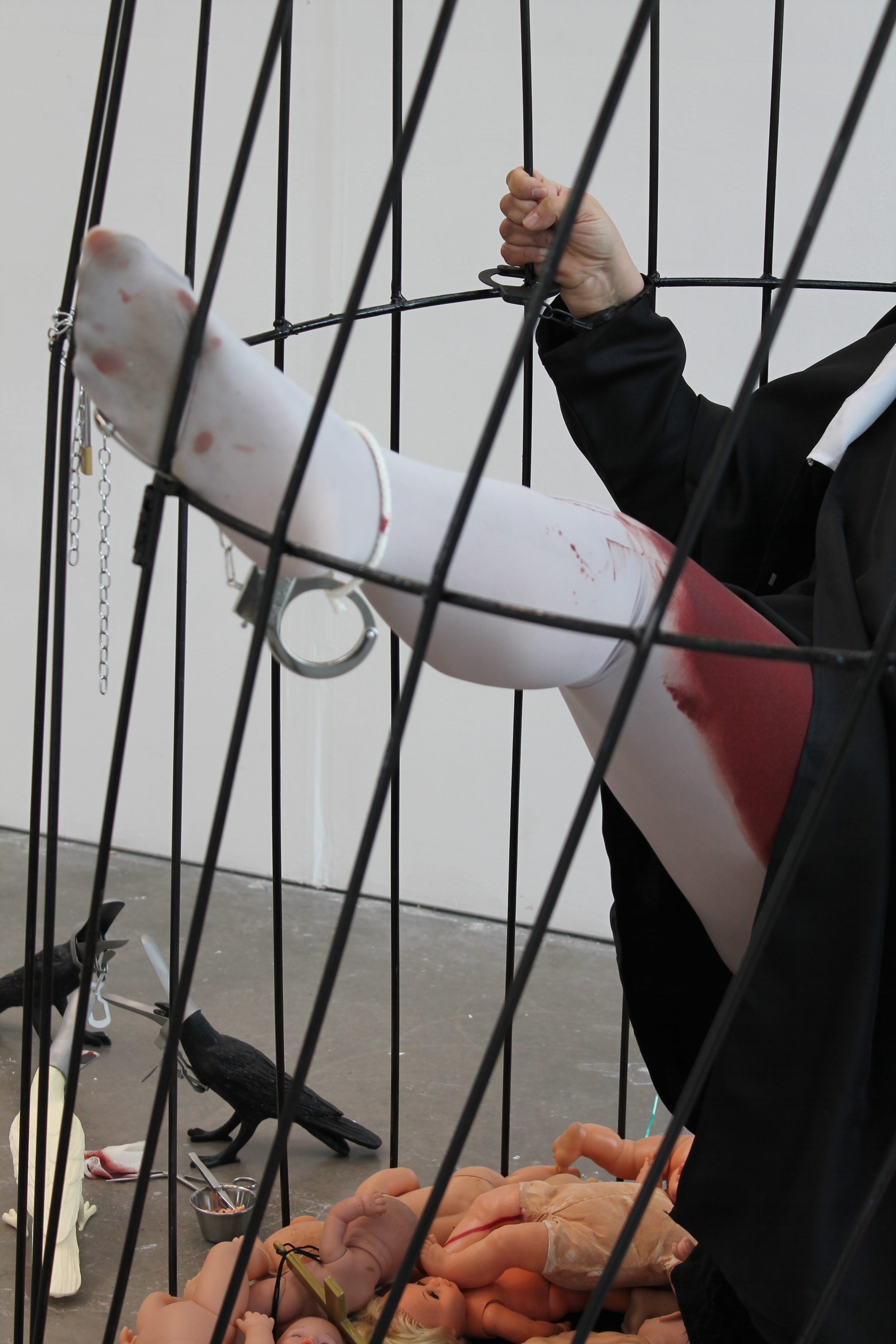 A leg in white tights with blood at the top of the thigh is raised. The person is a metal cage and the rest of the body is out of the frame aart from a hand holding one of the bars. The floor of the cage is covered in baby dolls and plastic crows sit
