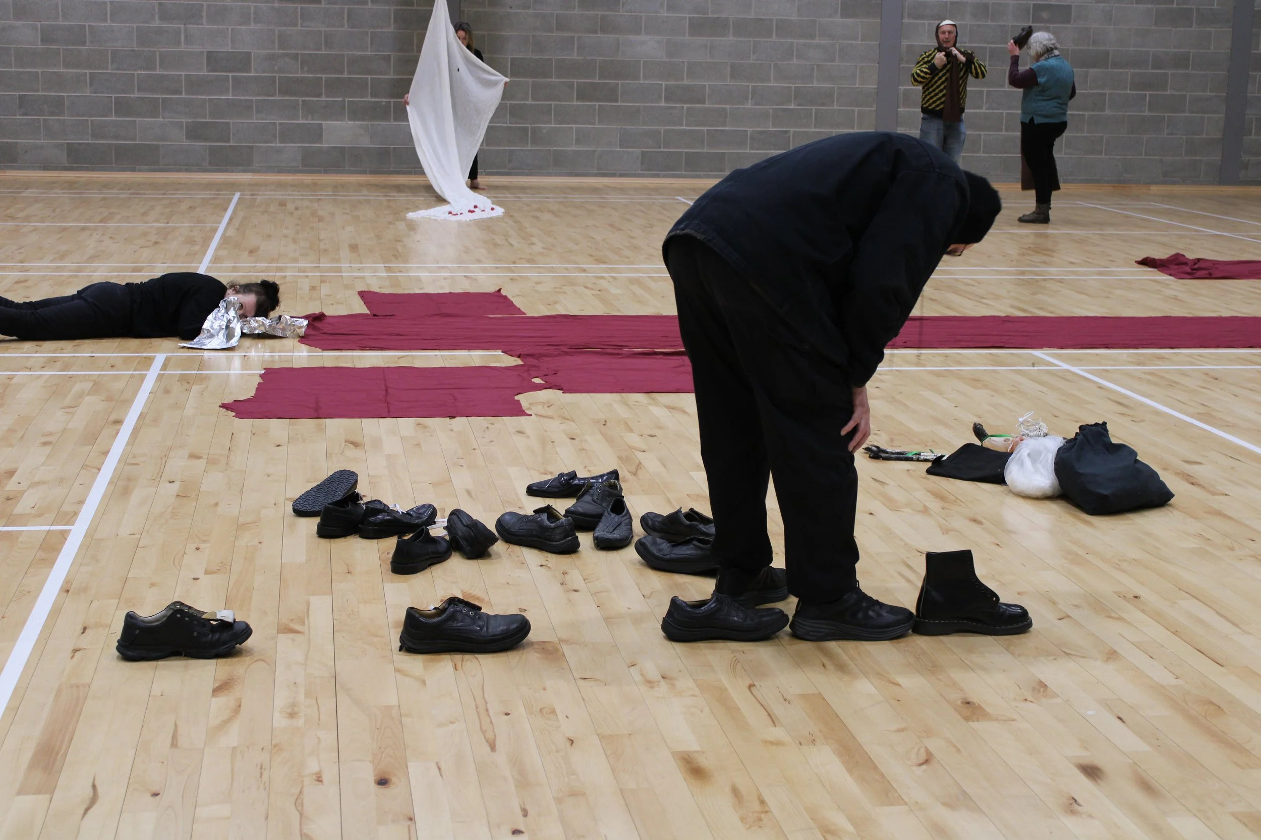 In a sports hall, front centre is man in black bent over looking at 8 pairs of black shoes on the floor, large red swathes of fabric laid on the floor behind him, someone lies on the floor, left, with tin foil covering the face, person in background