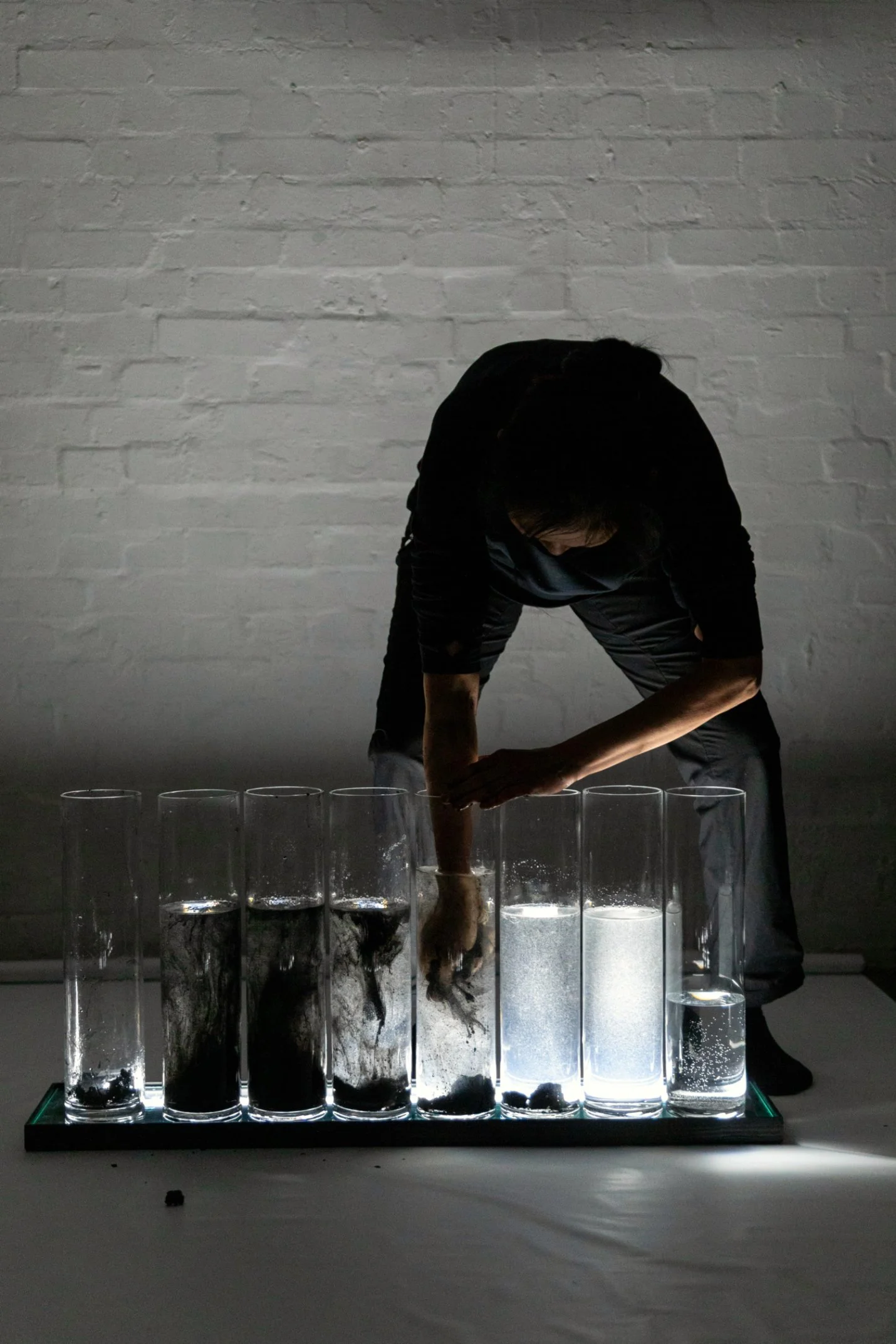 Person dressed n black with a row of 8 knee height round  glass vessels three quarter filled with liquid, some have dirt and roots in them. The person is leaned over with one hand in the liquid in one jar. Soft lighting, white brick wall and floor
