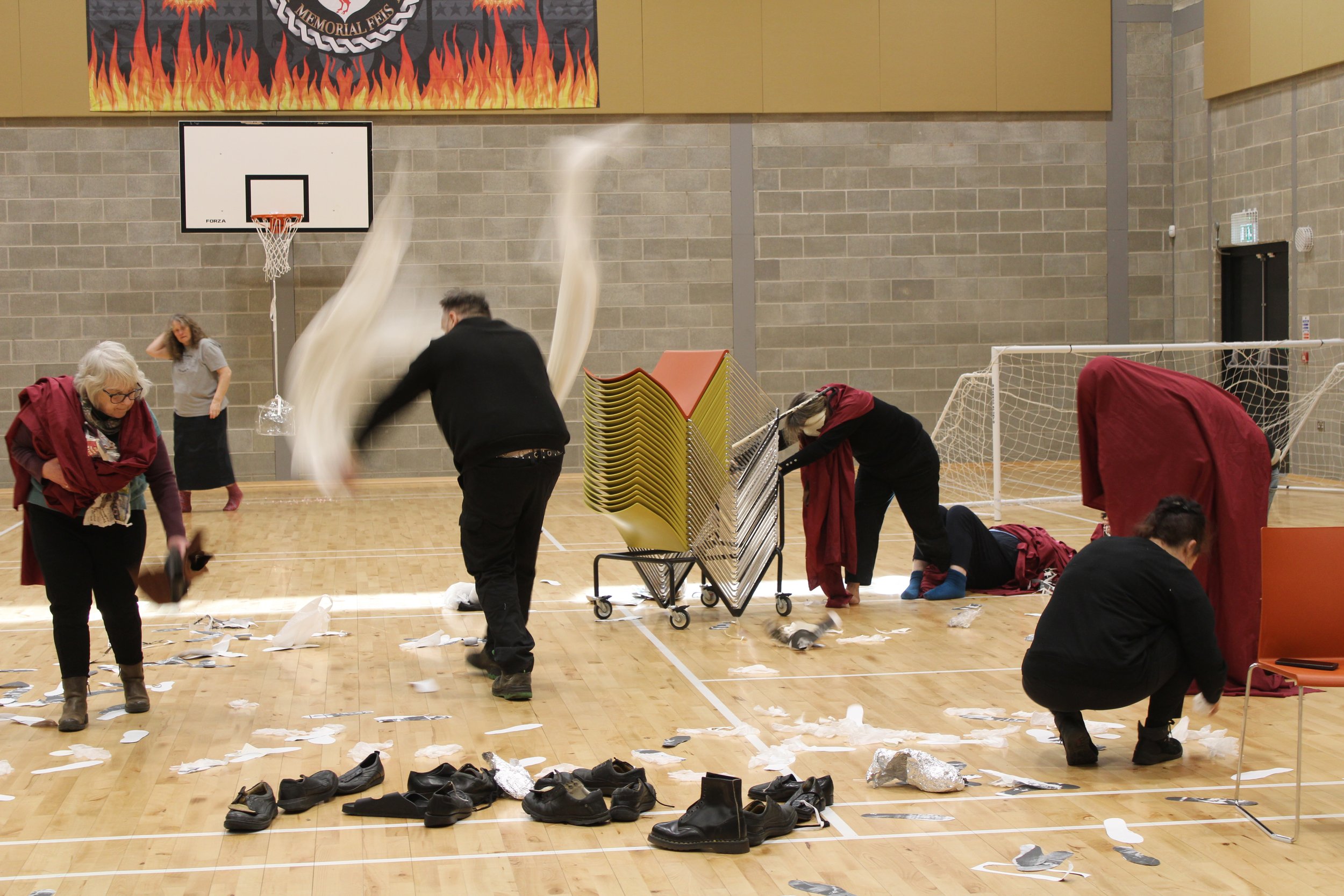 people in a sports hall, woman swathed in red fabric carrying a baf, man in black waving long white fabric strips in the air, person pushing stacked trolley of chairs, lots of black shoes and white paper on the floor, person in black hunched on floor