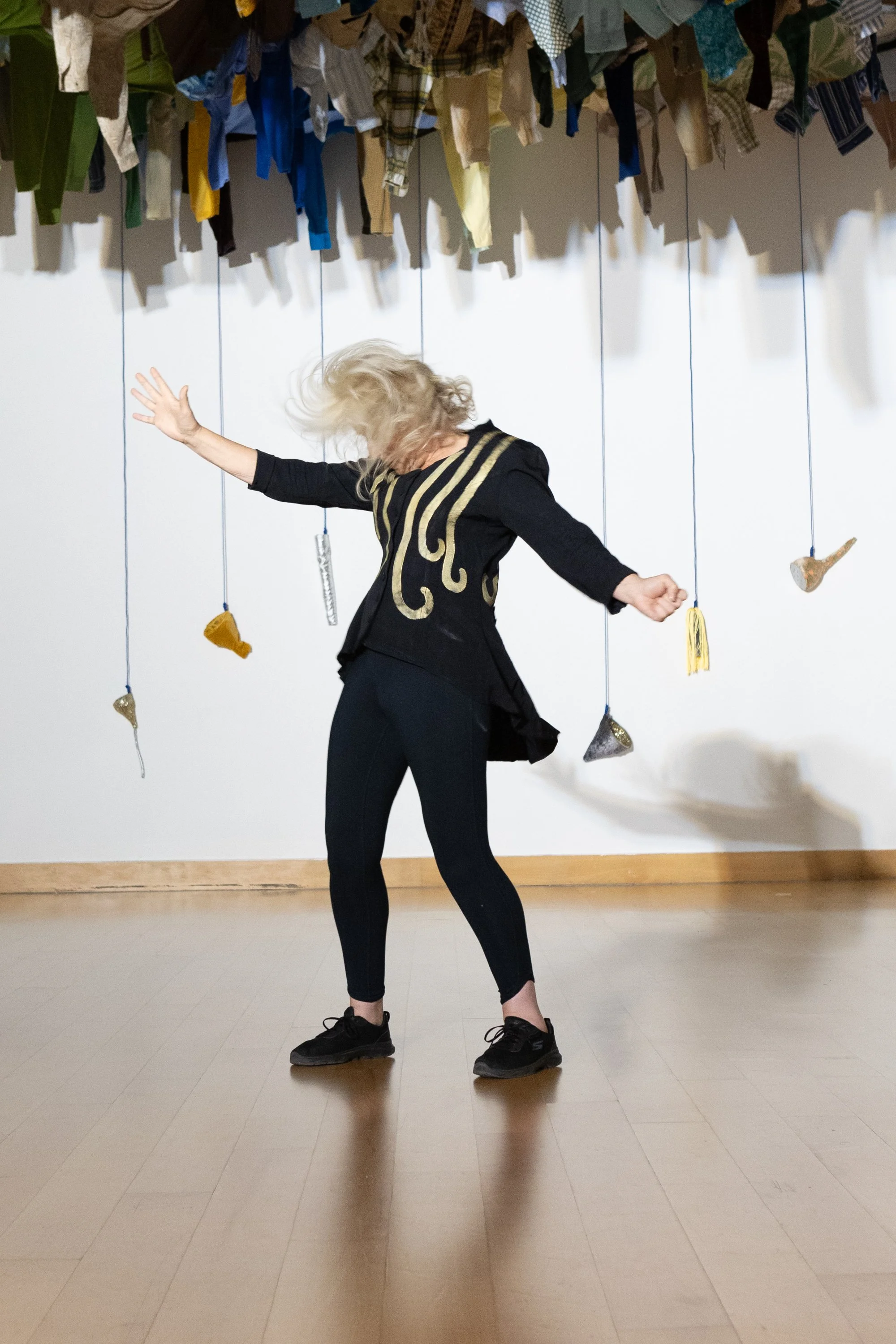 blonde woman swishing her heair, wearing black trousers and top with gold lines dow the front, in a white space with  clothes hung above and cups like vessels hung behind