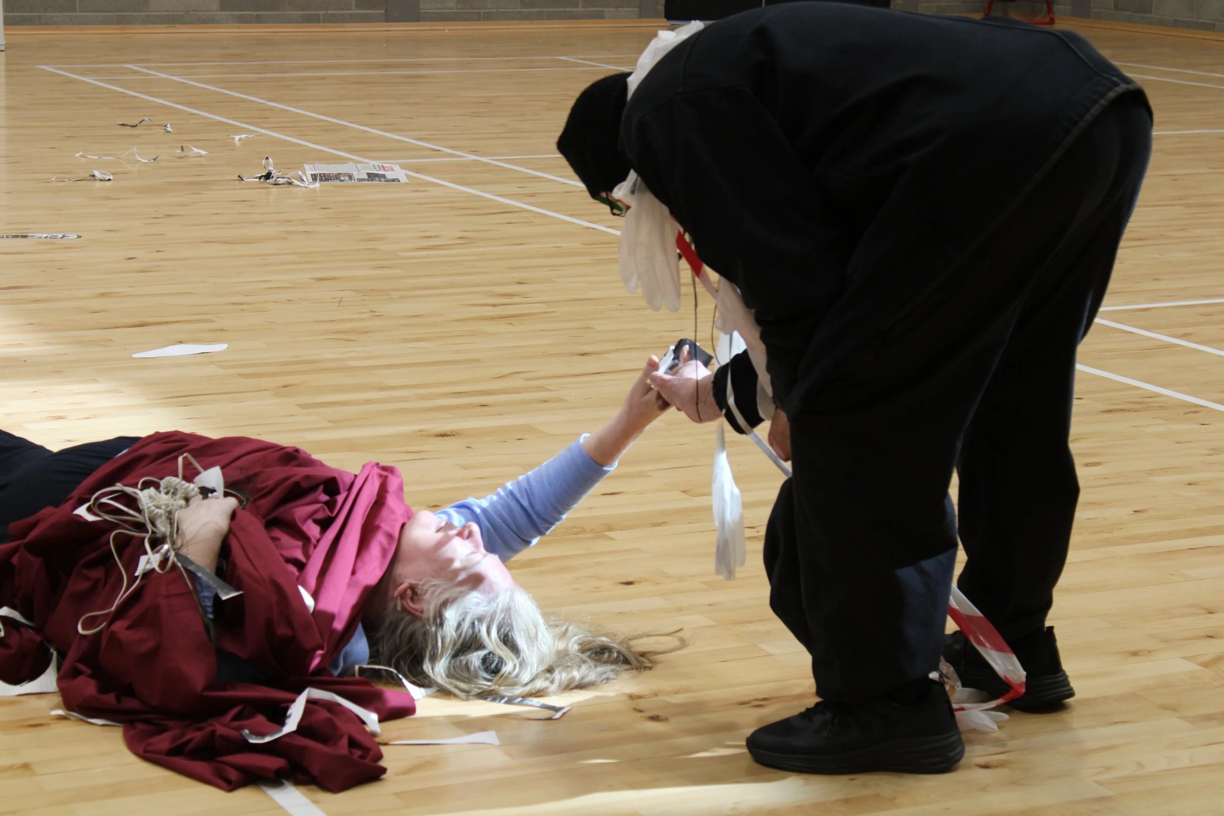 person lying on wooden sports hall floor swathed in burgundy fabric with right arm held out, man in black hunched over woman placing something in her hand