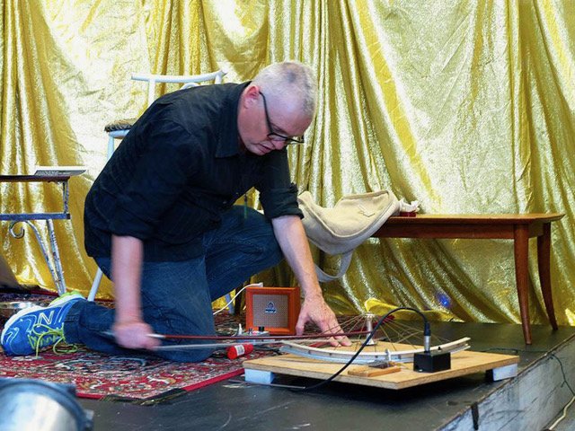 man kneeling playing a horizontal bicycle wheel connected to pick ups on a wooden board with a violin bow. Gold curtains hanging behind him. small table behind him.