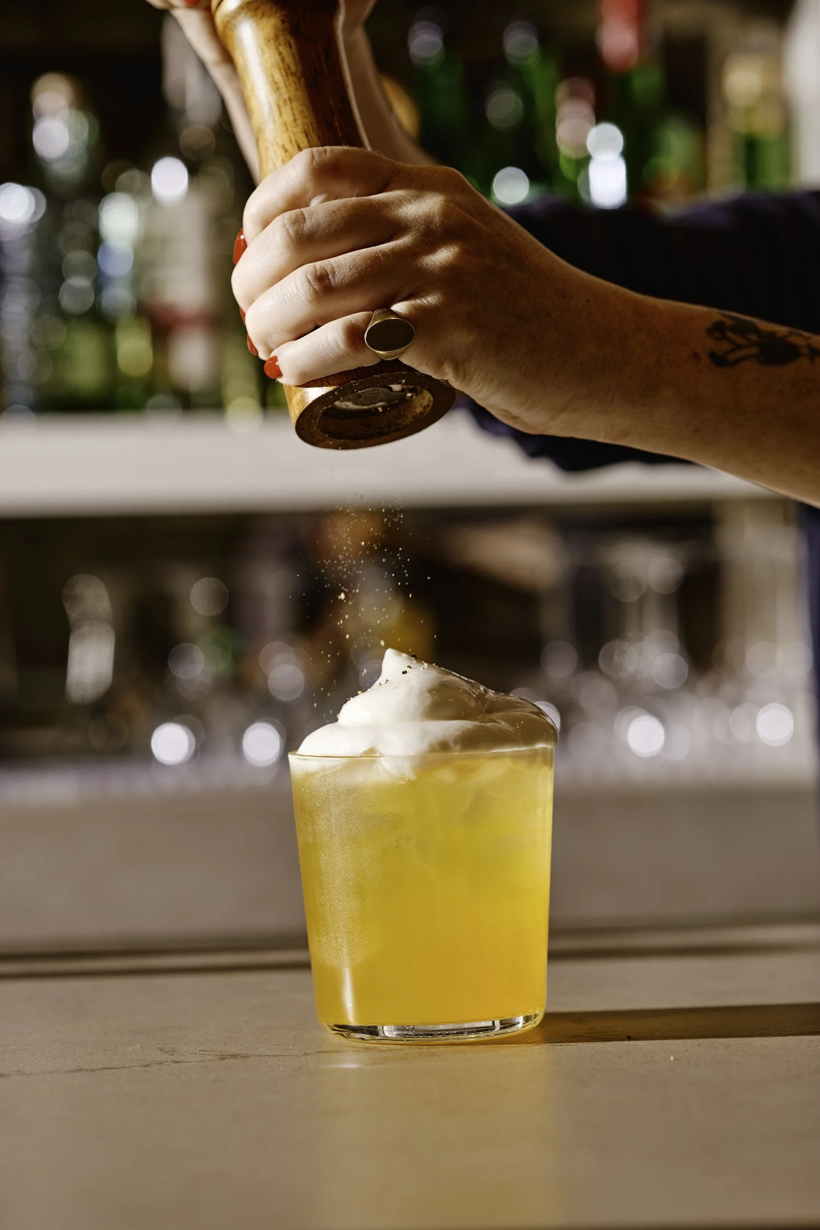 A bartender is sprinkling a spice or salt into a yellow cocktail with foam on top on a bar counter, with a background of liquor bottles.