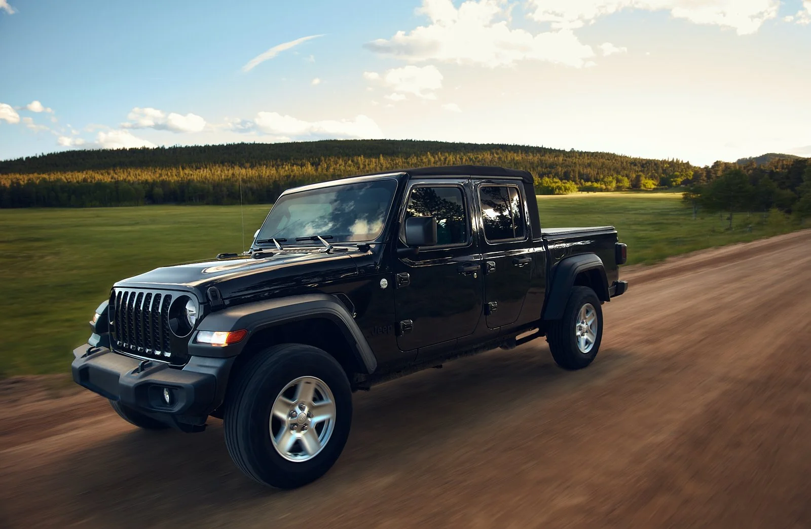 A black Jeep Gladiator pickup truck driving on a dirt road through a green field with trees and hills in the background, under a partly cloudy sky.