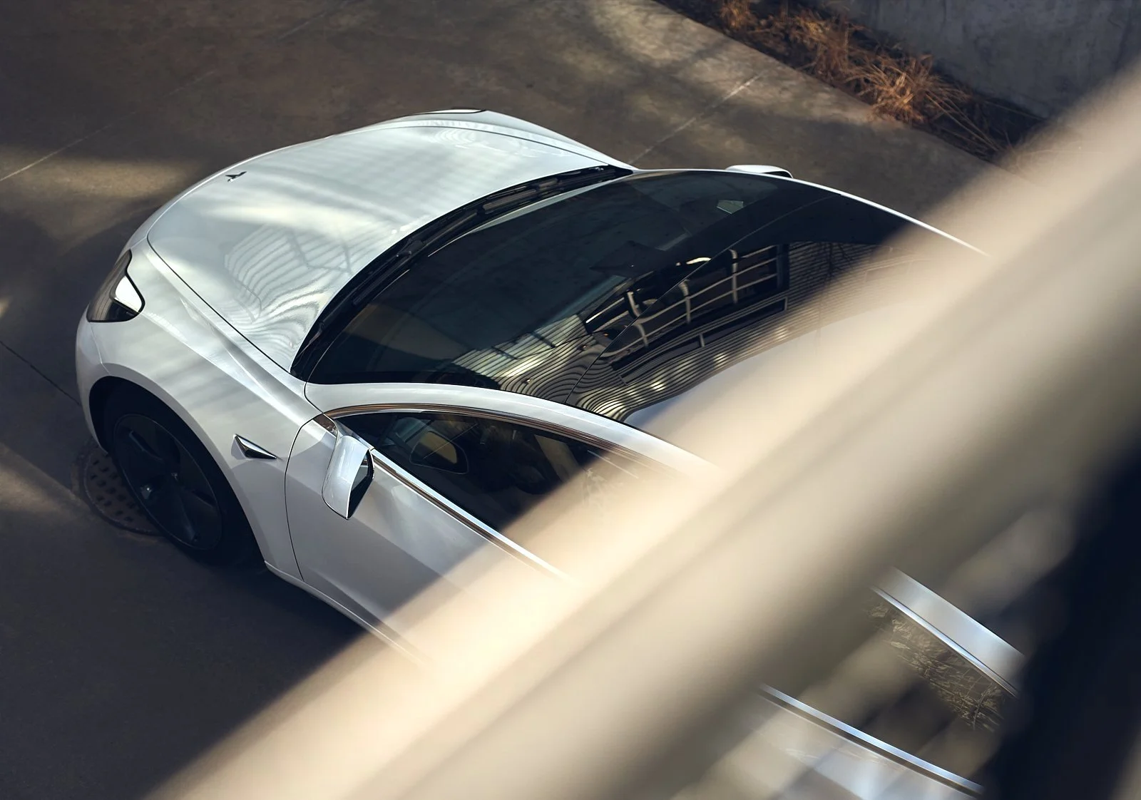 Top-down view of a white Tesla car parked on a street, partially obscured by a blurred railing or fence in the foreground.