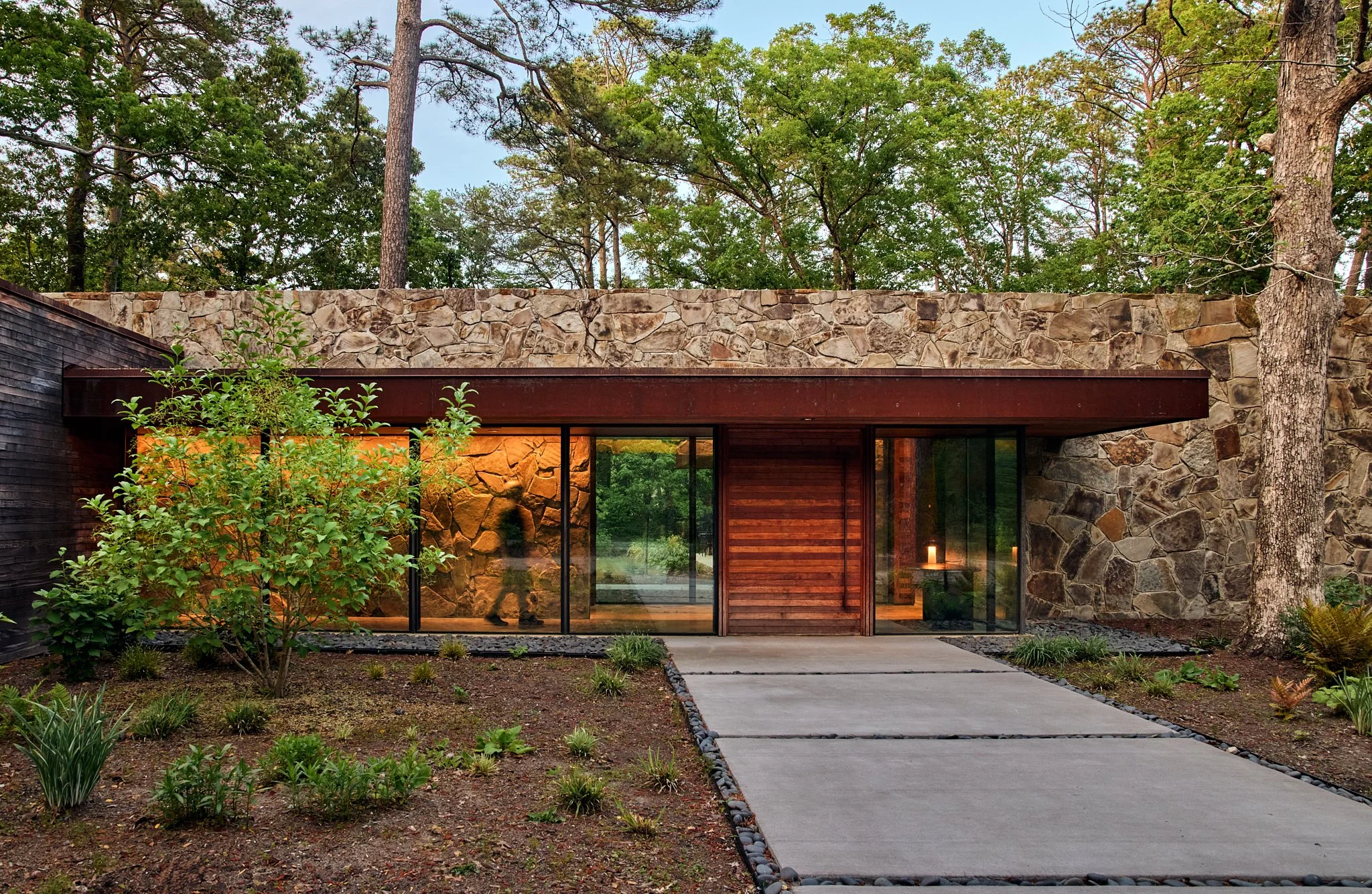 Exterior architectural photograph of a contemporary stone and wood residence set within a forest landscape. The low horizontal roofline, corten steel detailing, and expansive glass walls define a modern mountain home aesthetic. Warm interior lighting
