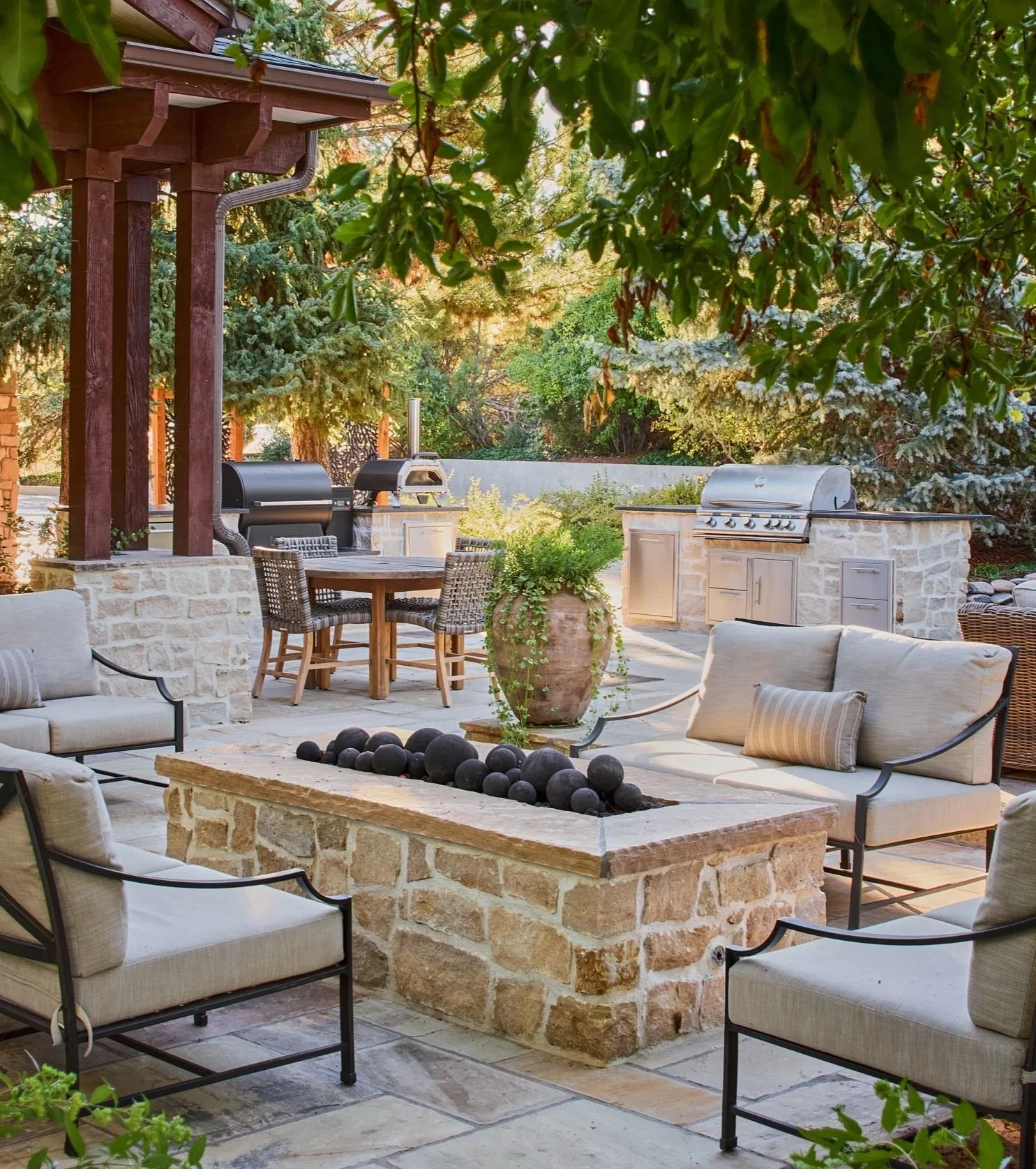 Outdoor patio area with cushioned chairs centered around a stone and brick firepit, trees and greenery in the background, and an outdoor kitchen with grills and a dining table.