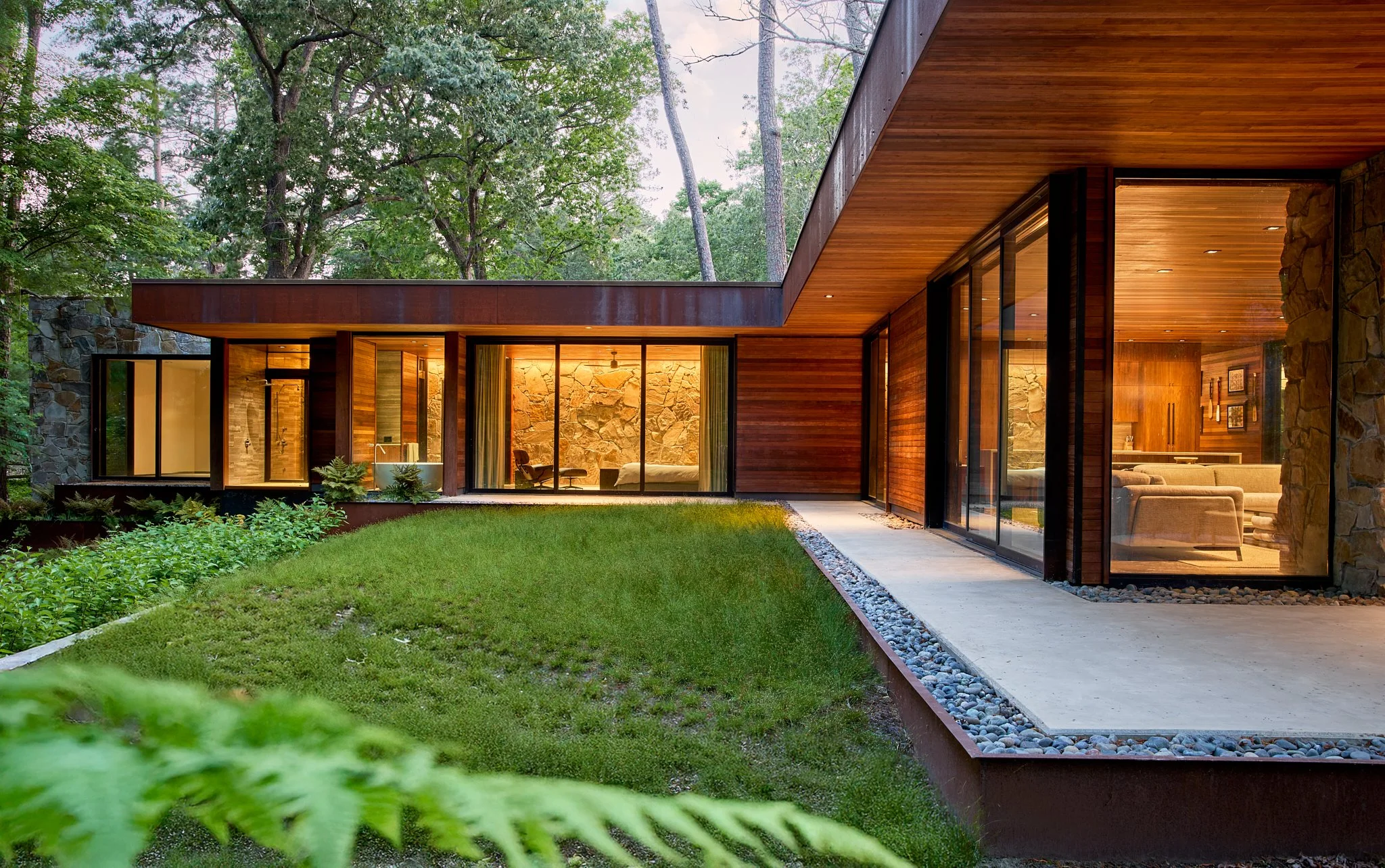 Exteriors Architectural photography Modern house with large glass windows, wooden exterior, stone accents, surrounded by trees and greenery, shown during evening with interior lights on.