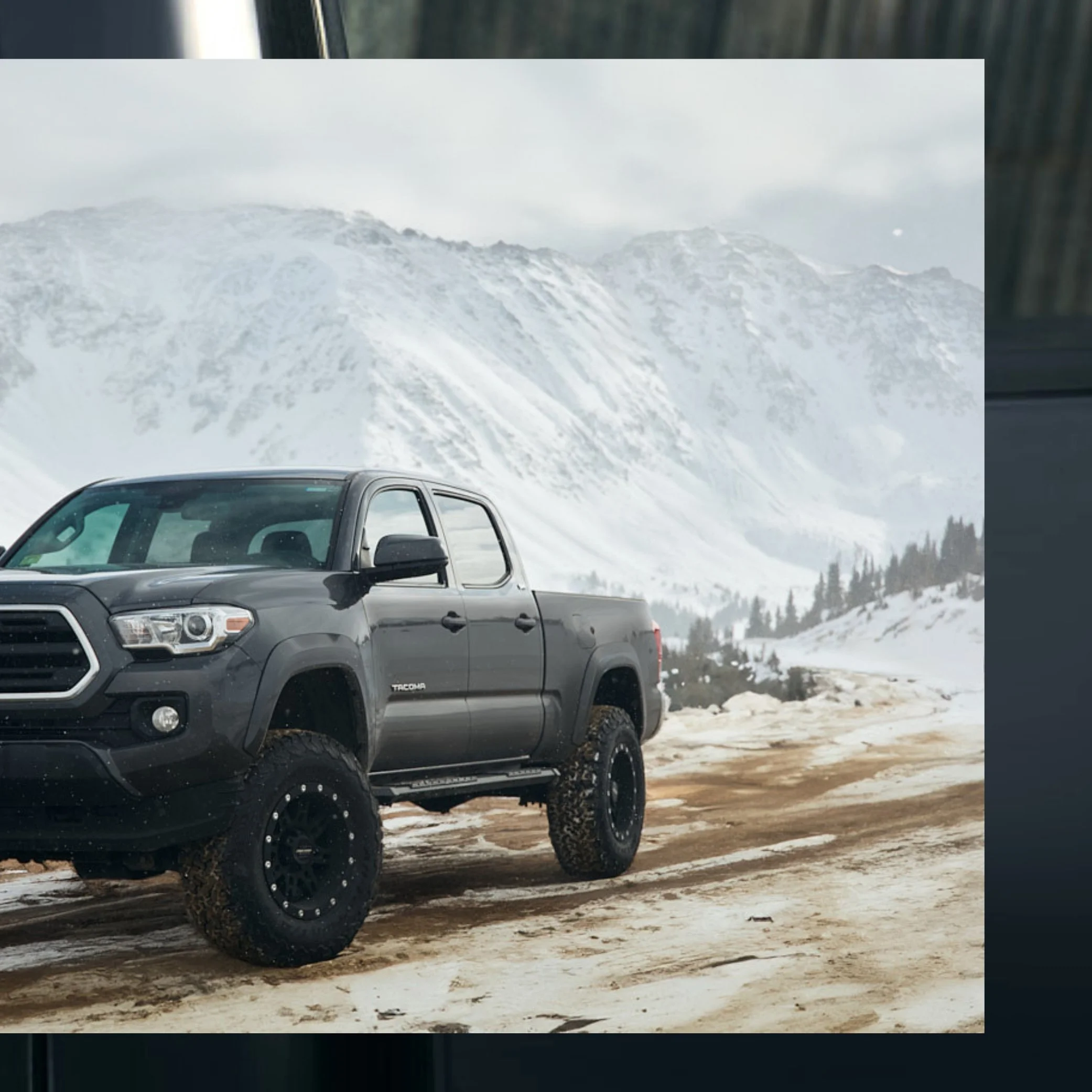 A black Toyota Tacoma pickup truck driving on a snowy mountain dirt road with snow-covered peaks and trees in the background.