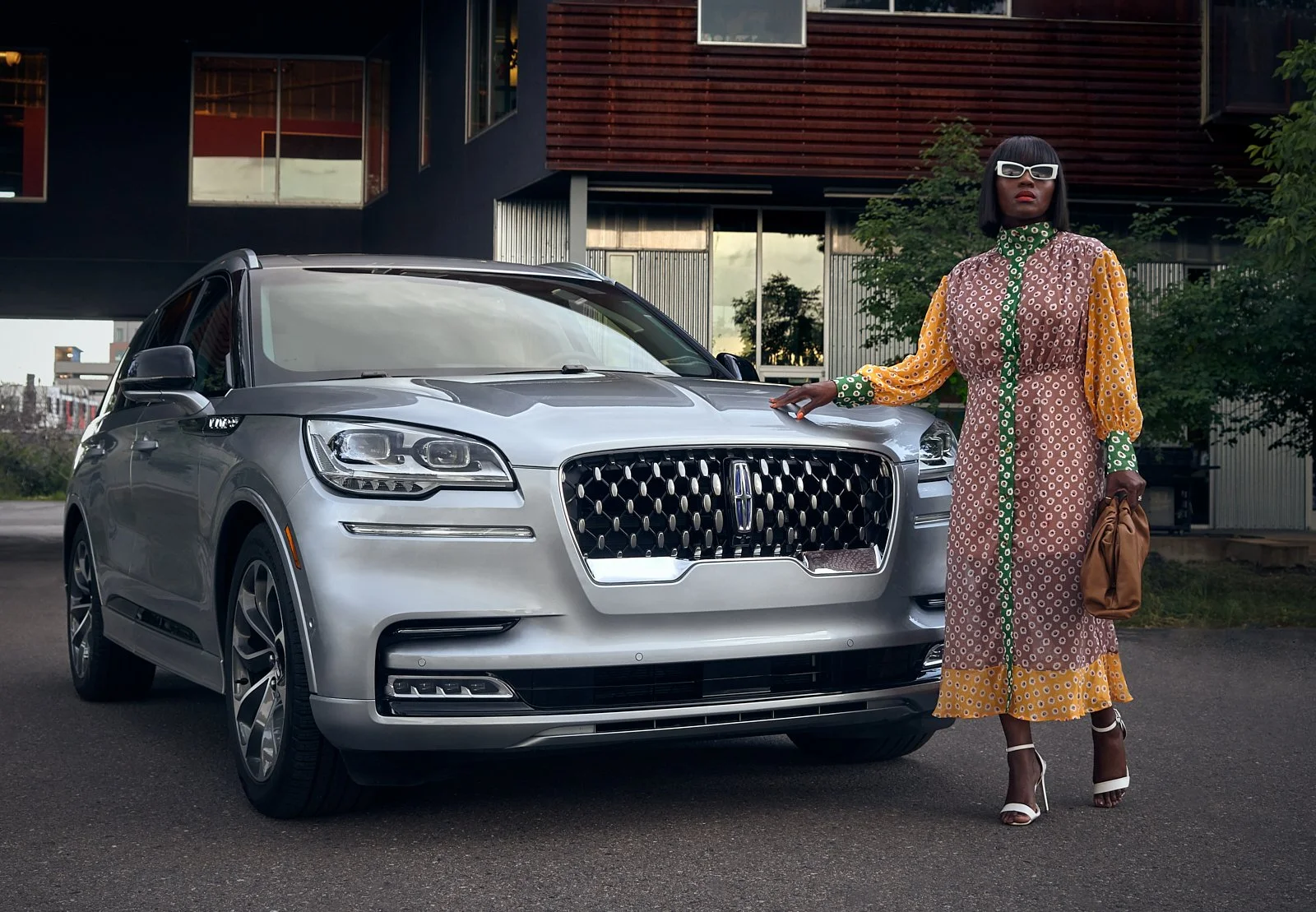 A woman with dark hair, wearing a colorful patterned dress, white sunglasses, and high heels, standing next to a silver luxury SUV in an urban setting.