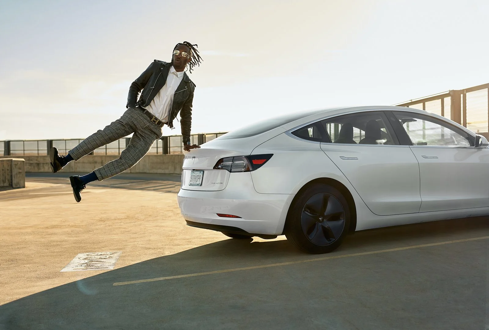 A man dressed in stylish clothing is leaning on a white Tesla car in a parking lot during sunset, with one leg in the air.