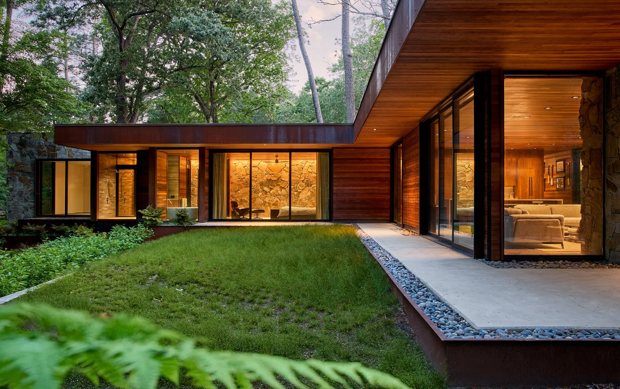 Twilight architectural photograph of a modern wood-and-stone residence designed to blend seamlessly with its natural surroundings. Featuring flat rooflines, floor-to-ceiling glass walls, and warm interior lighting, the home emphasizes indoor-outdoor 