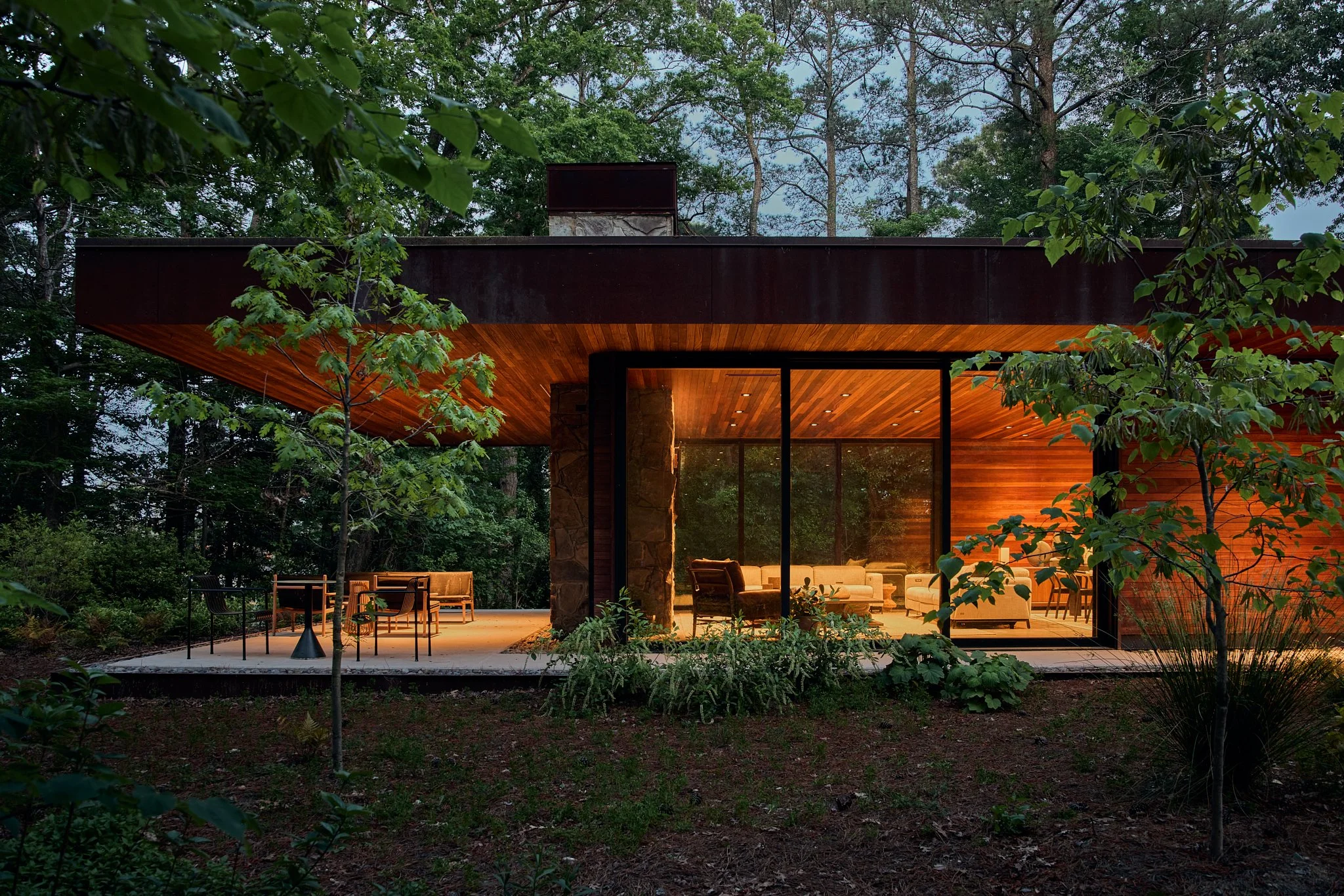 Twilight architectural photograph of a modern flat-roof residence featuring corten steel cladding, wood ceilings, stone detailing, and expansive glass walls. Warm interior lighting contrasts with the surrounding forest landscape, emphasizing indoor-o