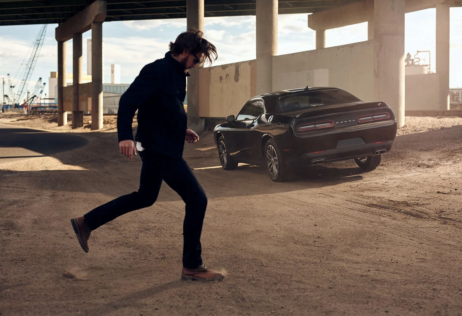 A man crossing a dirt lot under an overpass with a black Dodge Challenger parked behind him.