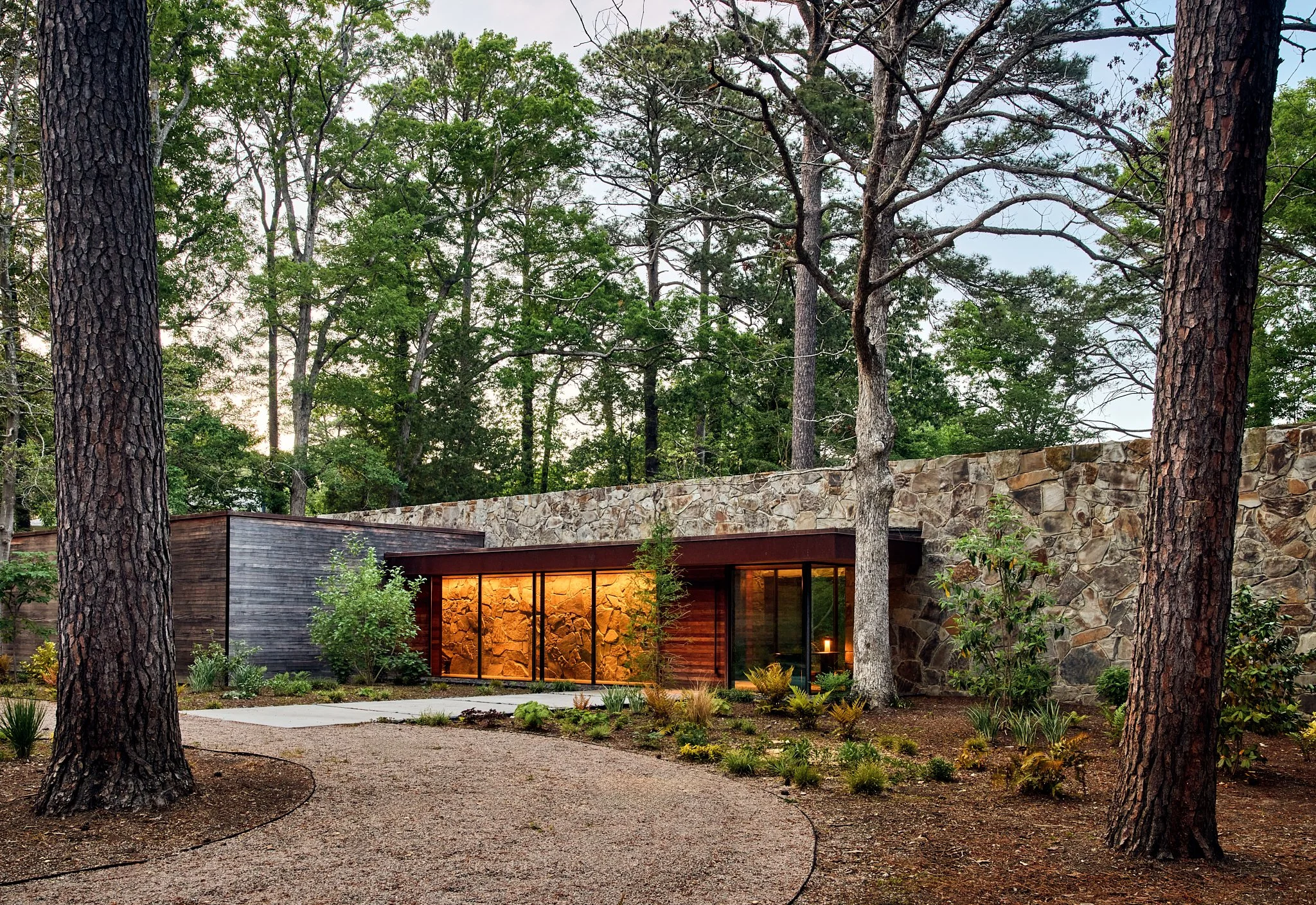 Wide exterior architectural photograph of a modern wood and stone residence set within a mature forest. The flat rooflines, expansive glass walls, and layered stone facade emphasize contemporary mountain home design and site-sensitive residential arc