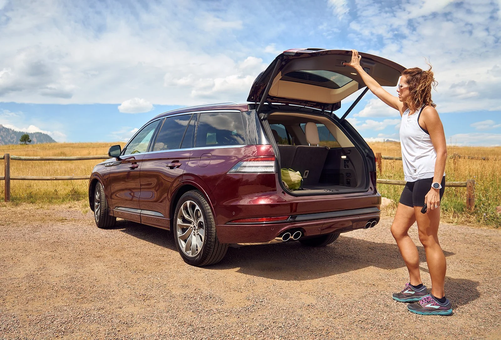 A woman standing outside a maroon SUV with the trunk open on a gravel area, with a grassy field and a partly cloudy sky in the background.