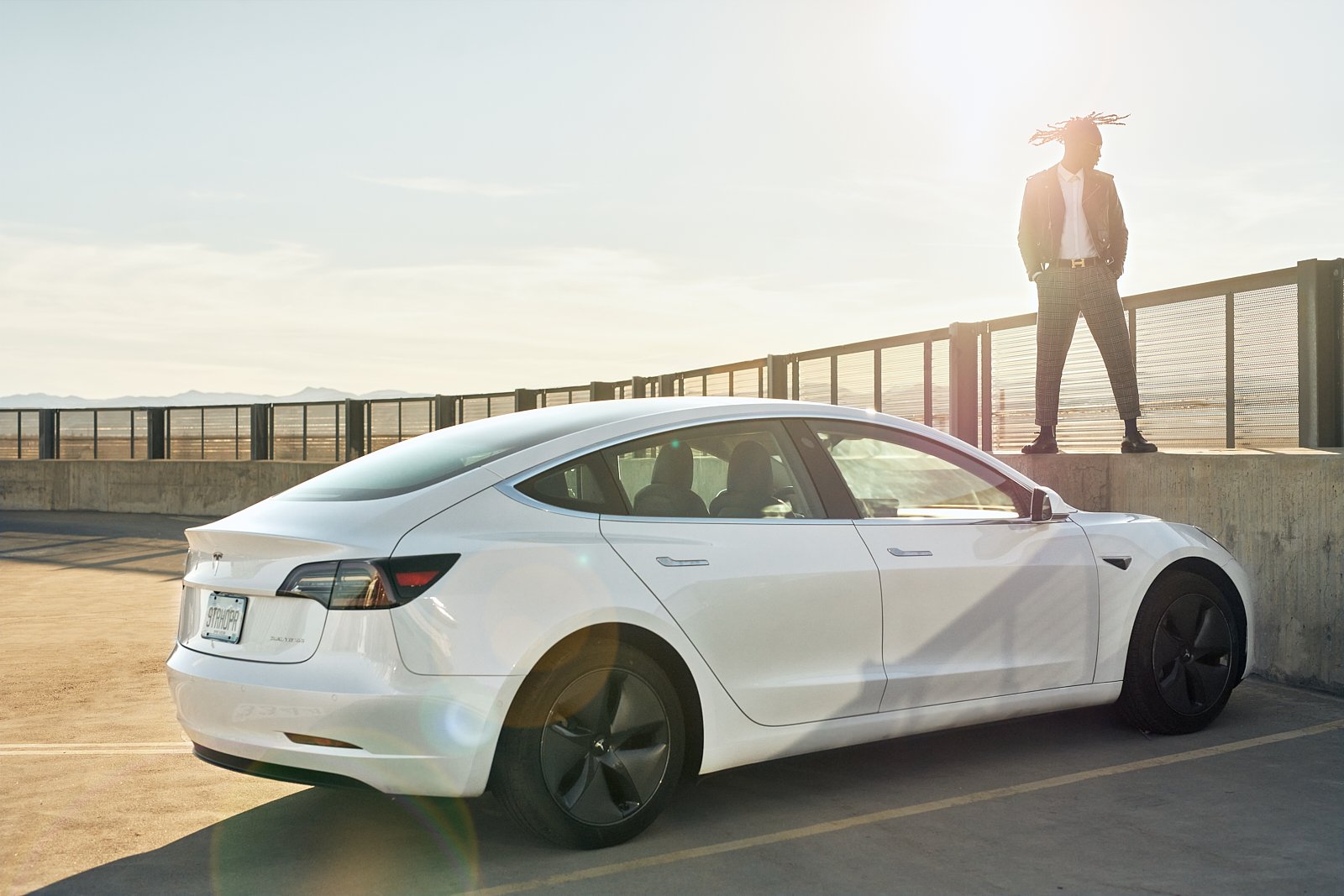 A white Tesla Model 3 parked on a rooftop parking lot during sunset. A woman with dreadlocks, dressed in a blazer and checkered pants, stands on the ledge looking into the distance.
