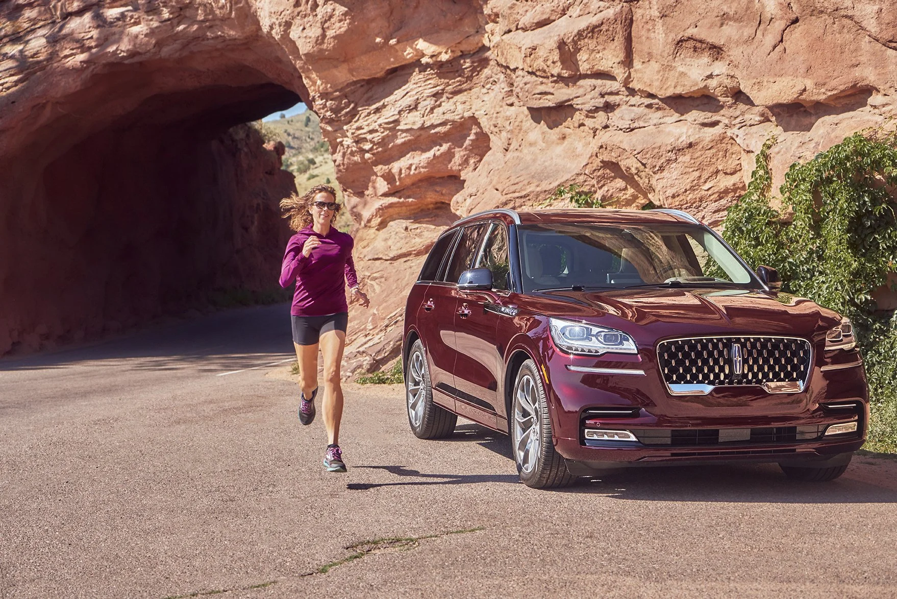 A woman jogging beside a shiny maroon SUV parked on a scenic road with red rock formations and a tunnel in the background.