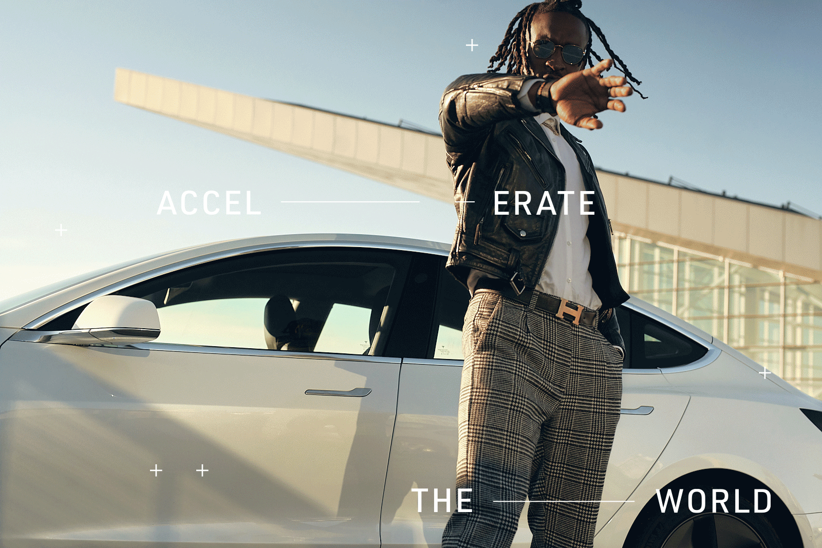 A man with dreadlocks wearing sunglasses, a leather jacket, and plaid pants stands next to a white car outdoors, with a modern building in the background, and the words 'Accelerate the world' overlaid.