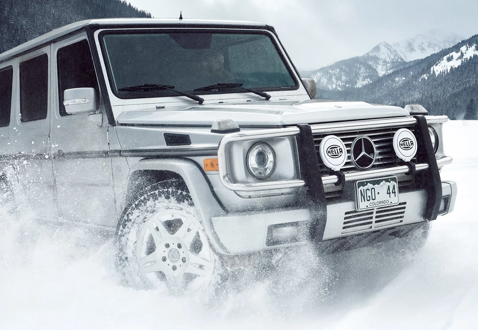 A silver Mercedes-Benz G-Class SUV driving through snow in a mountainous area with snow-covered trees and mountains in the background.