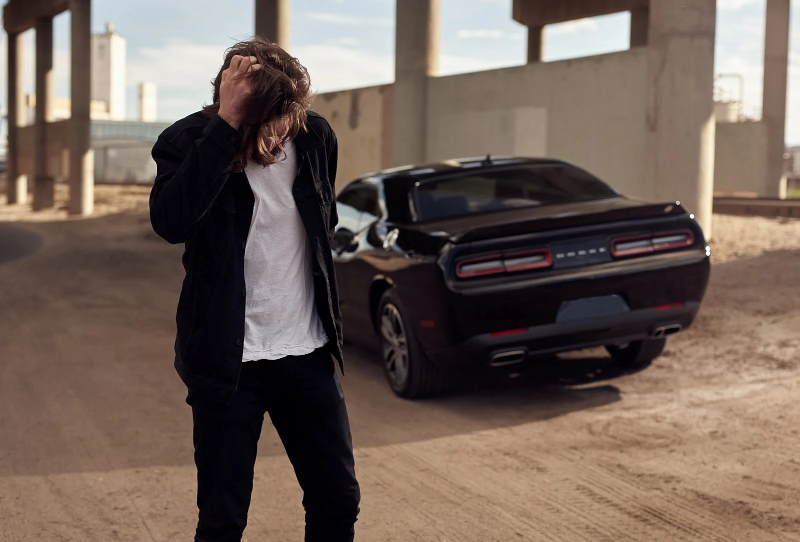 A person with long hair wearing a black jacket and white t-shirt standing next to a black Dodge Charger under a bridge with a distressed expression, hand on their head.