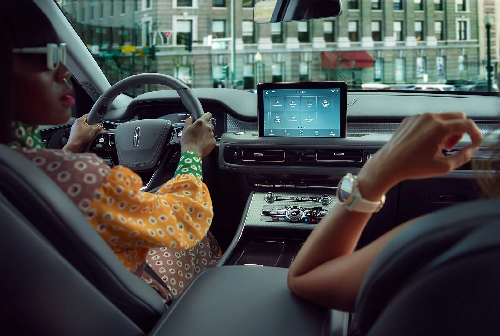 Interior of a modern car with a woman driving and another woman sitting next to her, looking at a large touchscreen display on the dashboard, with city buildings visible through the front windshield.