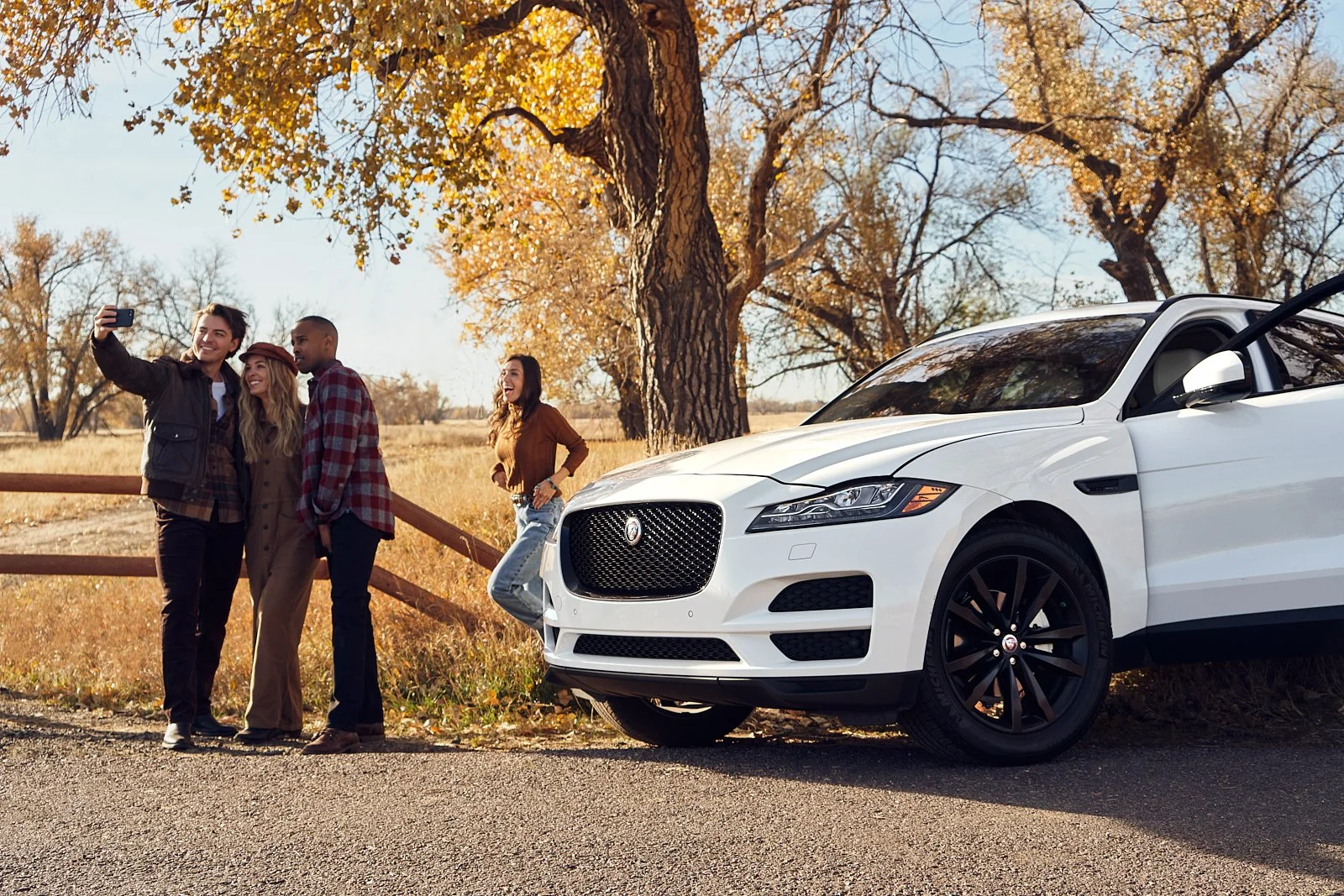 Four friends taking a selfie next to a white Jaguar car parked on a rural road with autumn trees in the background.
