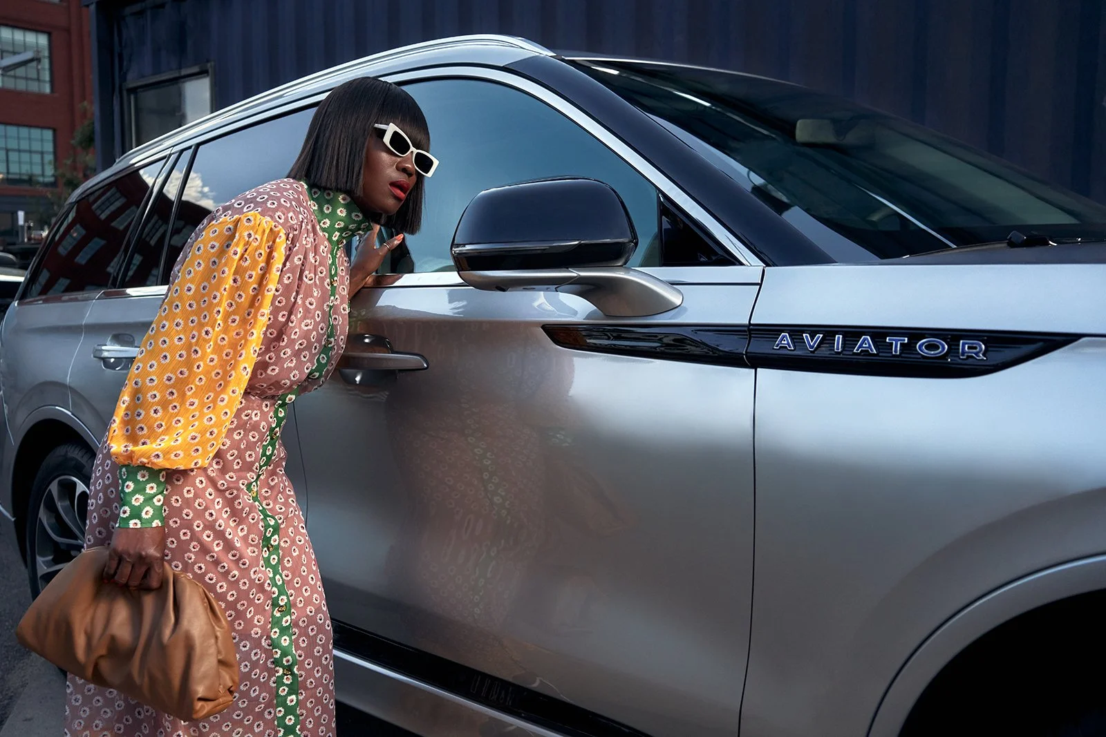 A woman with shoulder-length dark hair, wearing white sunglasses and a colorful patterned dress, is leaning toward a silver Land Rover Defender with the badge 'Aviator' on the side, holding a brown clutch bag, in an urban setting.