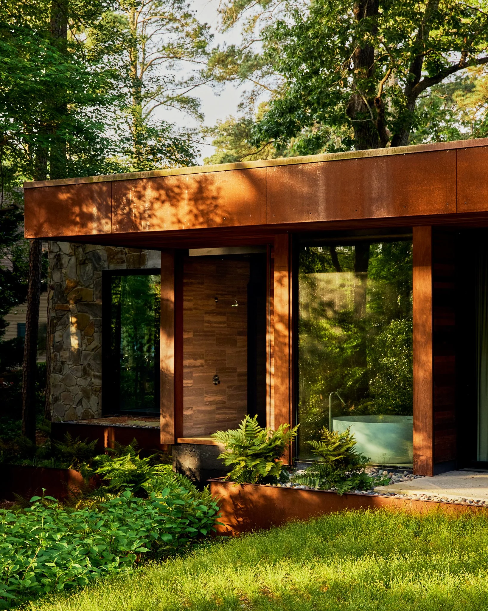 Architectural detail photograph of a modern residence featuring corten steel cladding, natural wood siding, and floor-to-ceiling glass set within a lush wooded landscape. Warm afternoon light highlights the layered materials and landscape integration