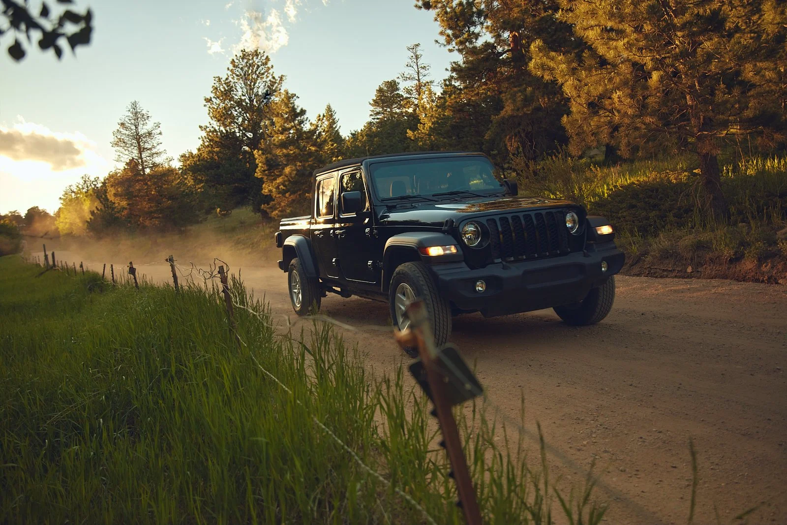 A black Jeep vehicle driving on a dirt road in a scenic outdoor setting with trees and grass, during sunset.