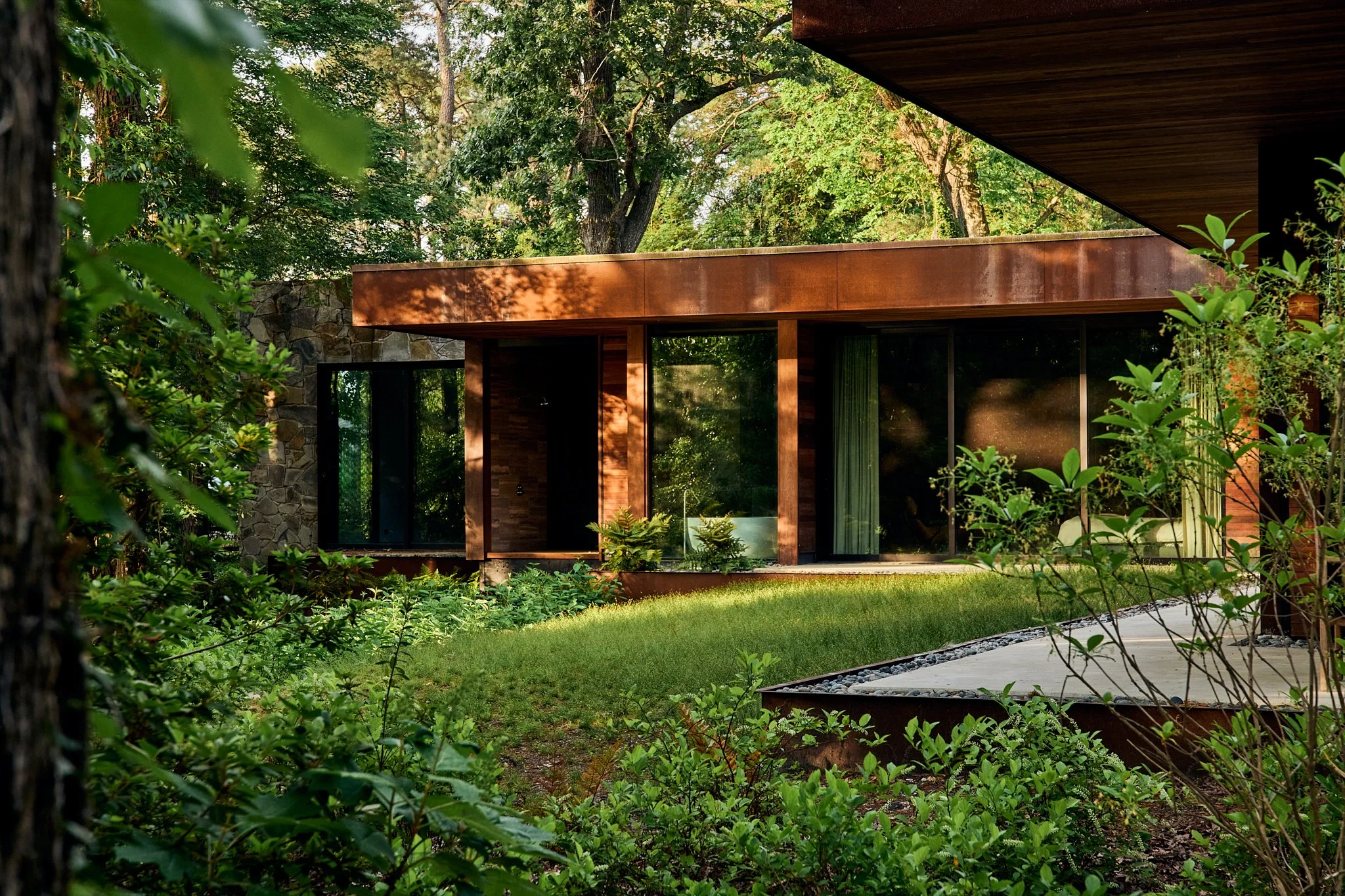 Architectural Photography of Modern house with large glass windows and rust-colored framing, surrounded by greenery and trees, with a lawn and pathway.