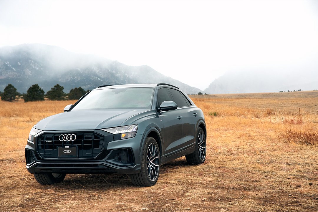 A gray Audi SUV parked in a dry, grassy field with mountains and cloudy sky in the background.