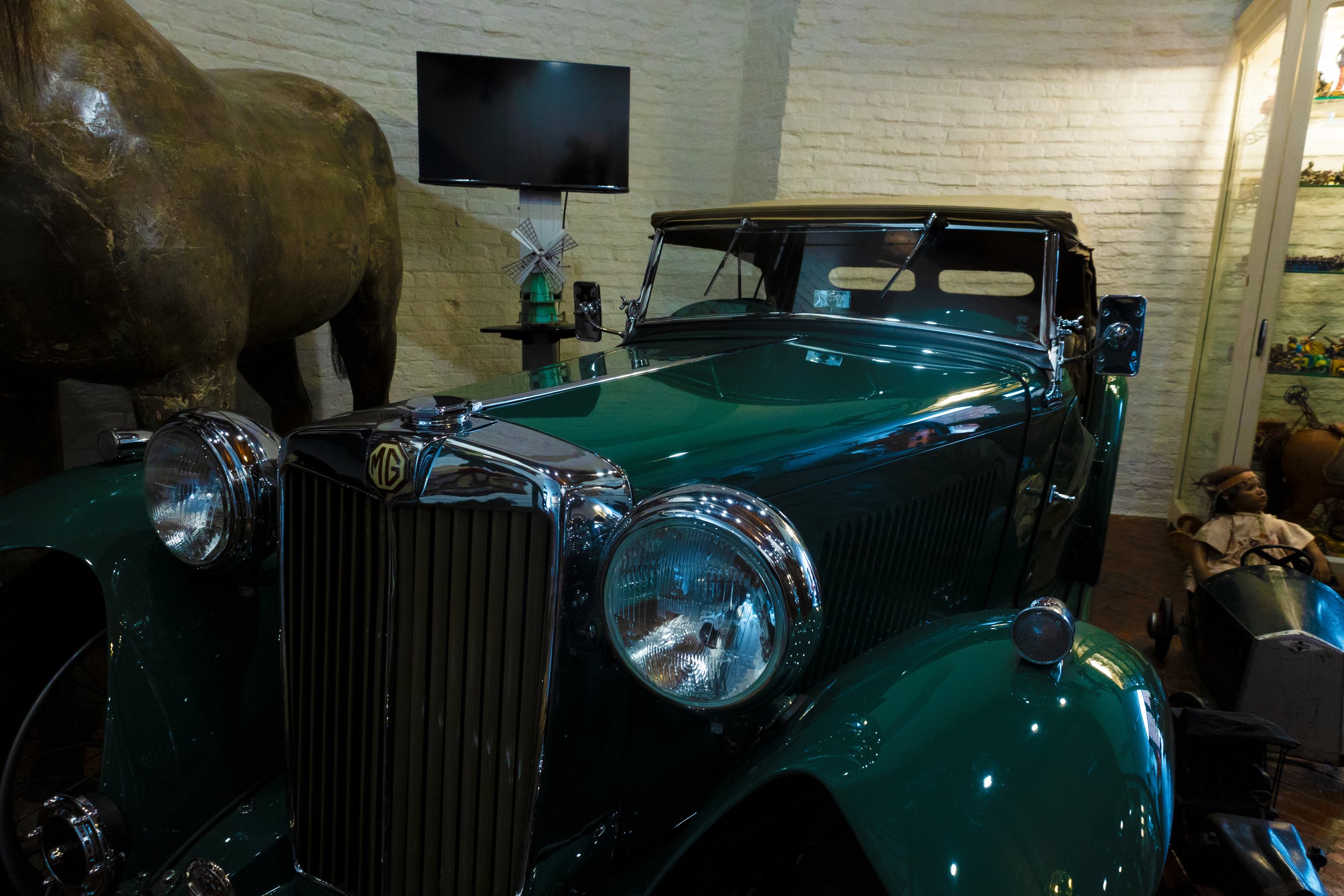 Groene vintage MG auto in een museum met een monitor, houten speelgoed en figurines op de achtergrond.