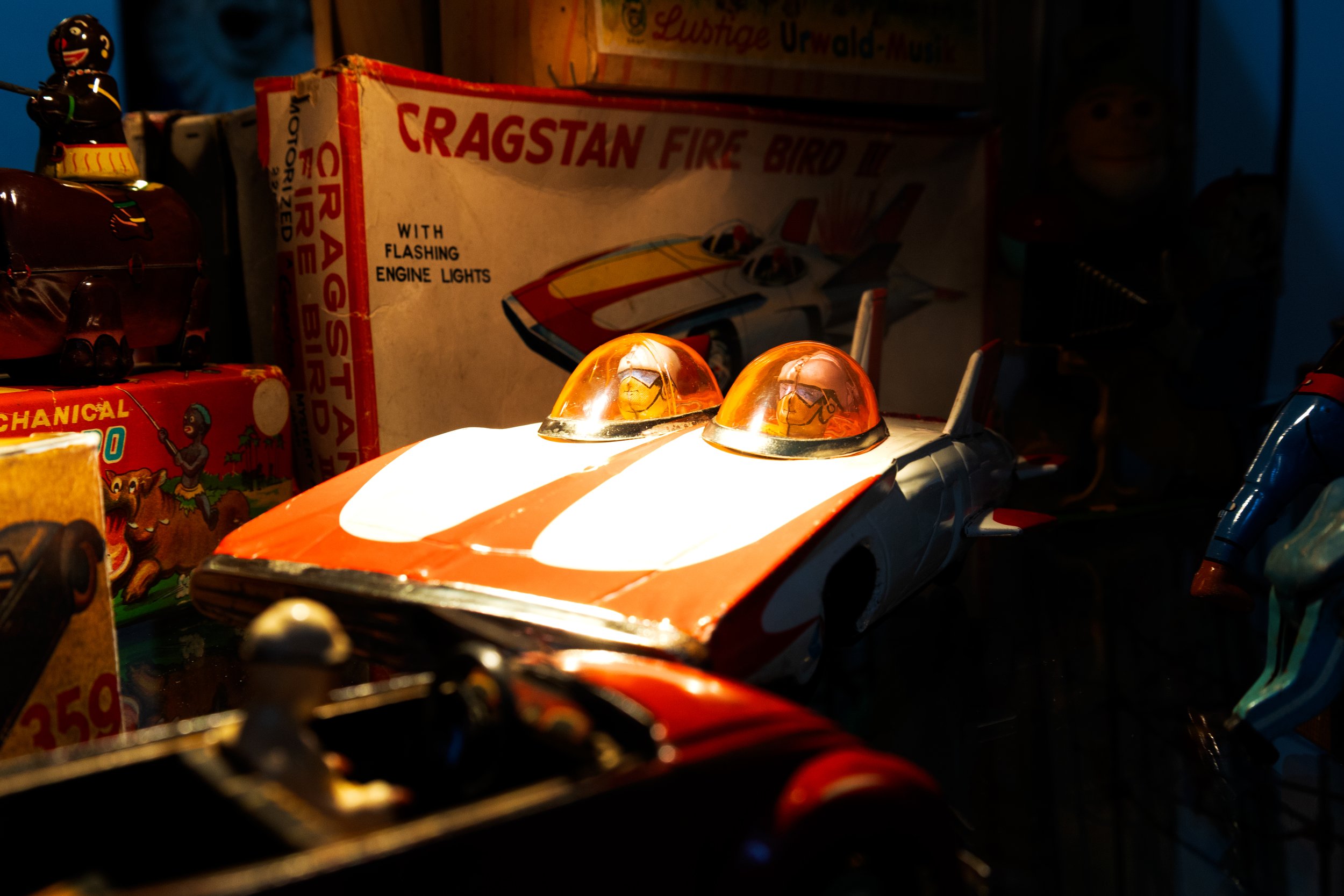 Een speelgoedauto met twee doorzichtige oranje cockpitopeningen, wit en rood gekleurd, omgeven door andere speelgoeditems en vintage speelgoedverpakkingen.