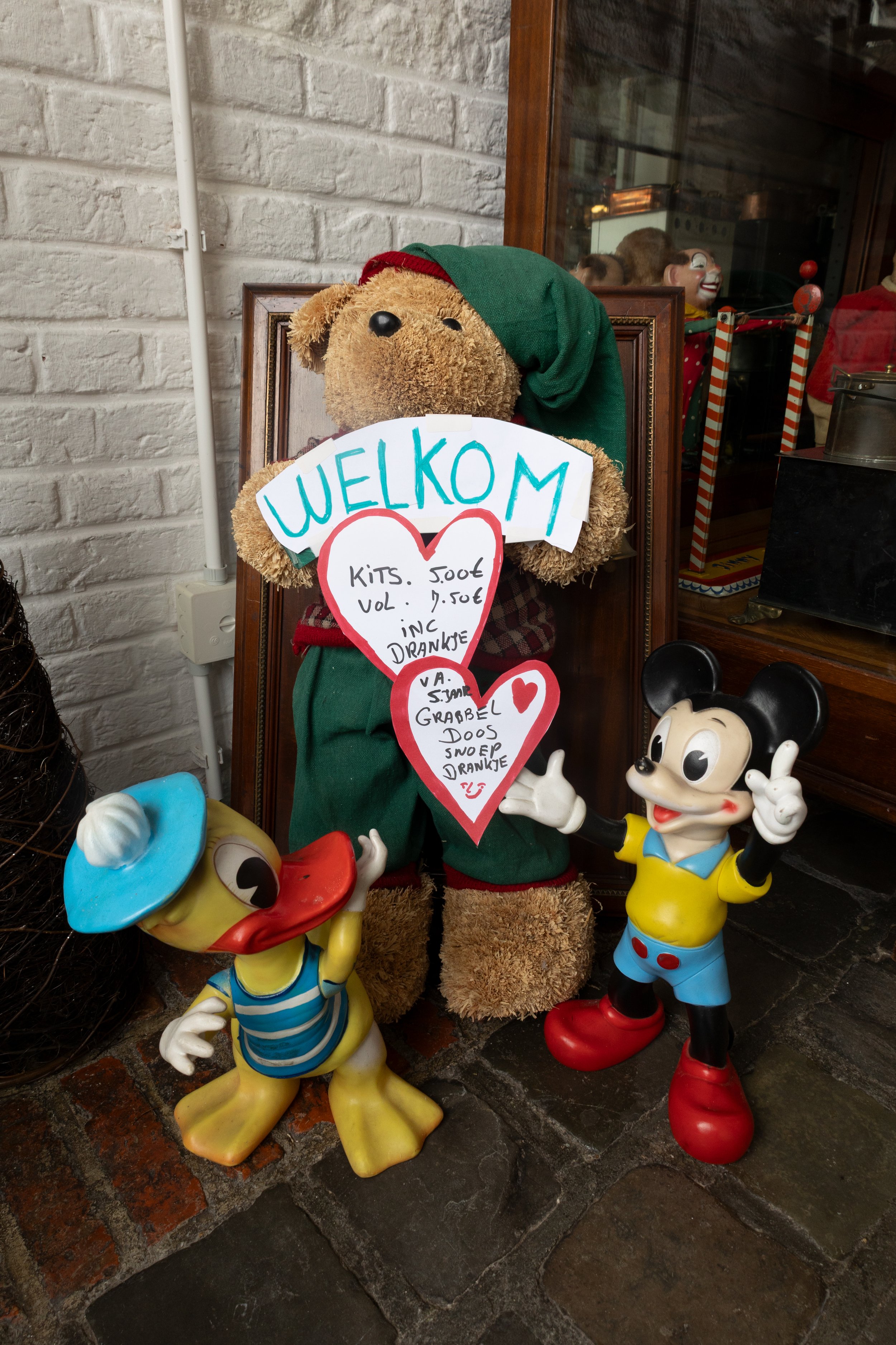 Decoratieve kerstfiguren, waaronder een teddybeer in een elfachtig pak en twee Disney-personages, Mickey Mouse en een eend, met een welkom bord en prijslijst in het Nederlands.